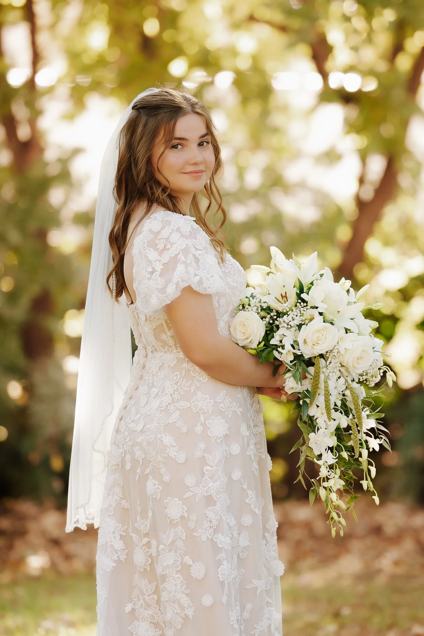 A bride in a white lace wedding dress holding a bouquet of white roses and lilies outdoors, with trees and sunlight in the background.