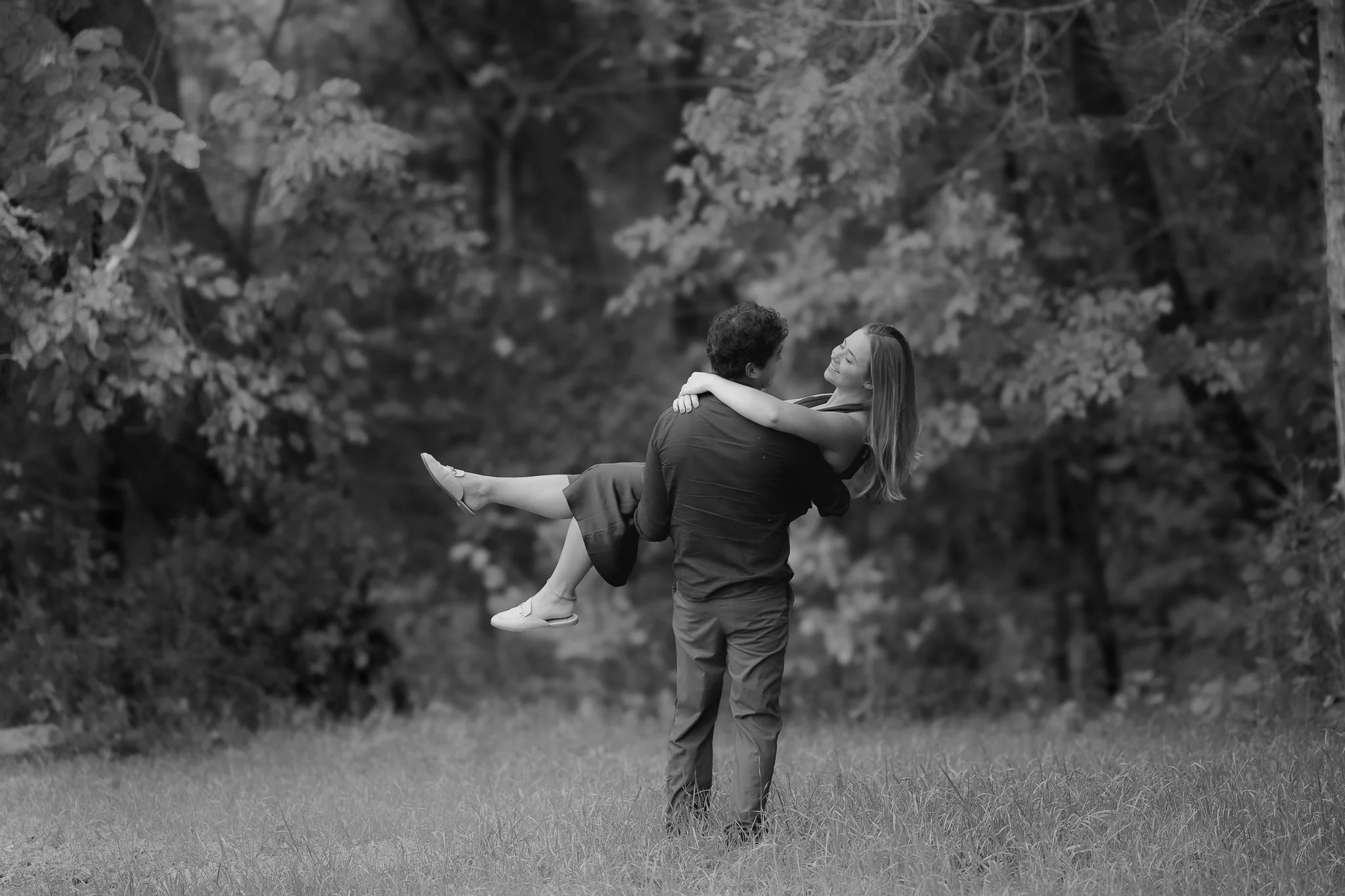 A couple in love, with the man holding the woman as she smiles and wraps her arms around his shoulders, standing on a grassy field with trees in the background.