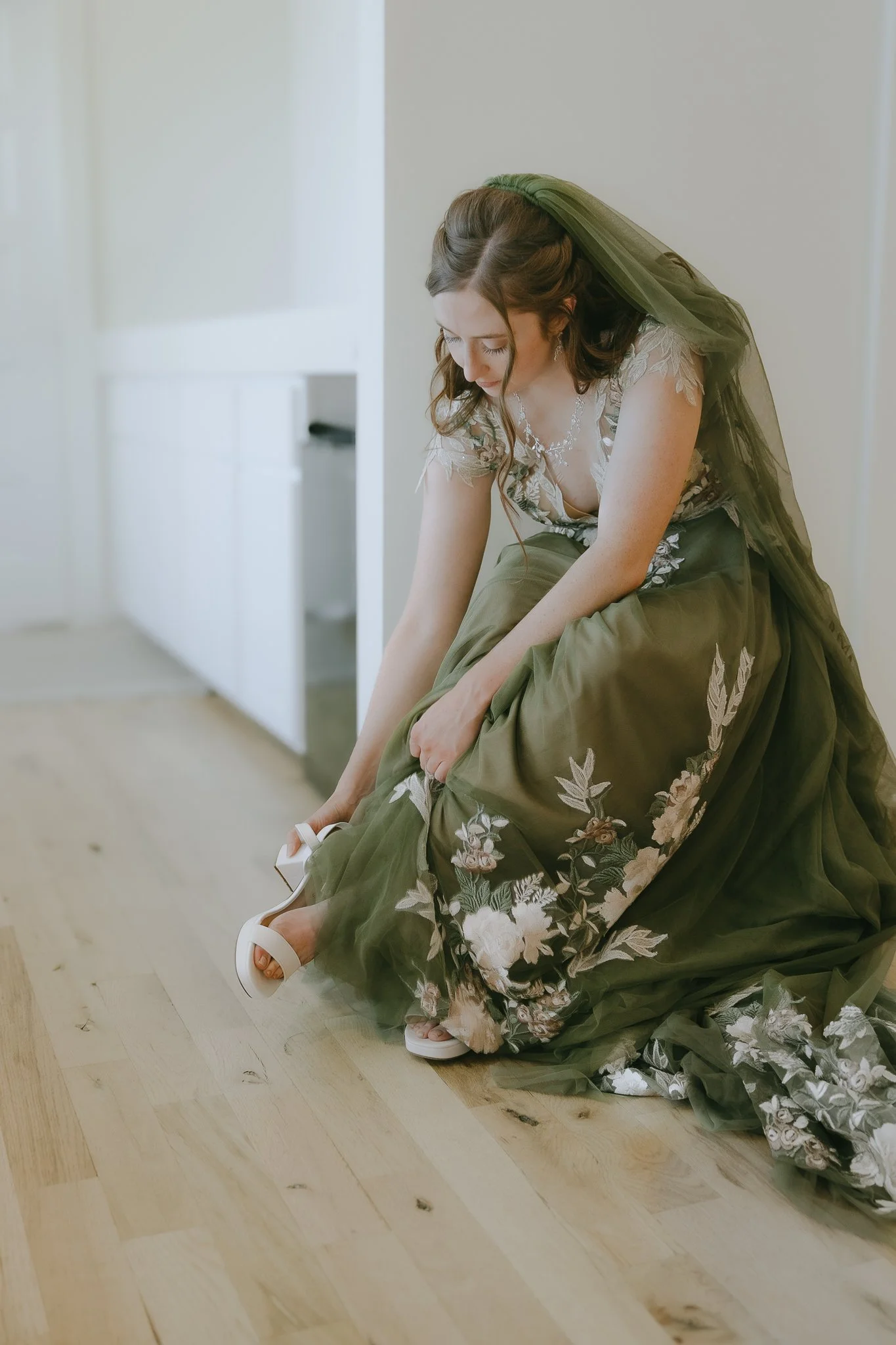 A bride in a green floral wedding dress putting on white high heels inside a bright room.