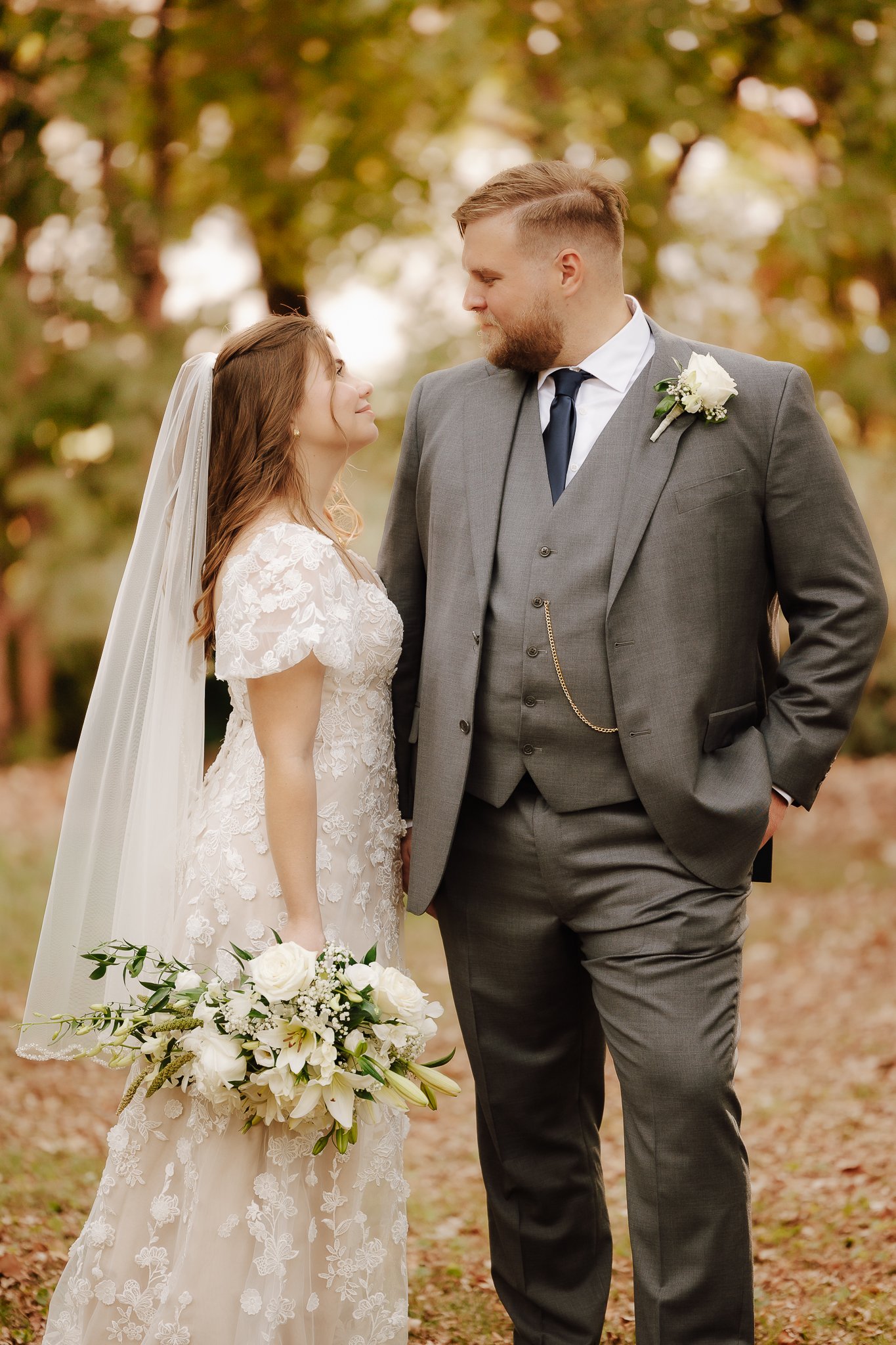 A bride and groom standing close together outdoors in autumn, gazing into each other's eyes, with trees and fallen leaves in the background.