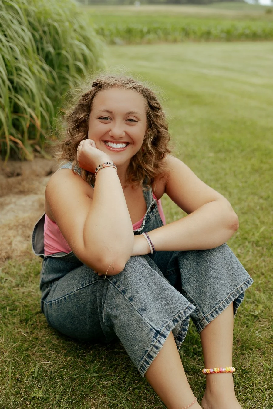 A young woman with curly hair smiling and sitting on grass near a pond, wearing denim overalls, pink top, and colorful bracelets.