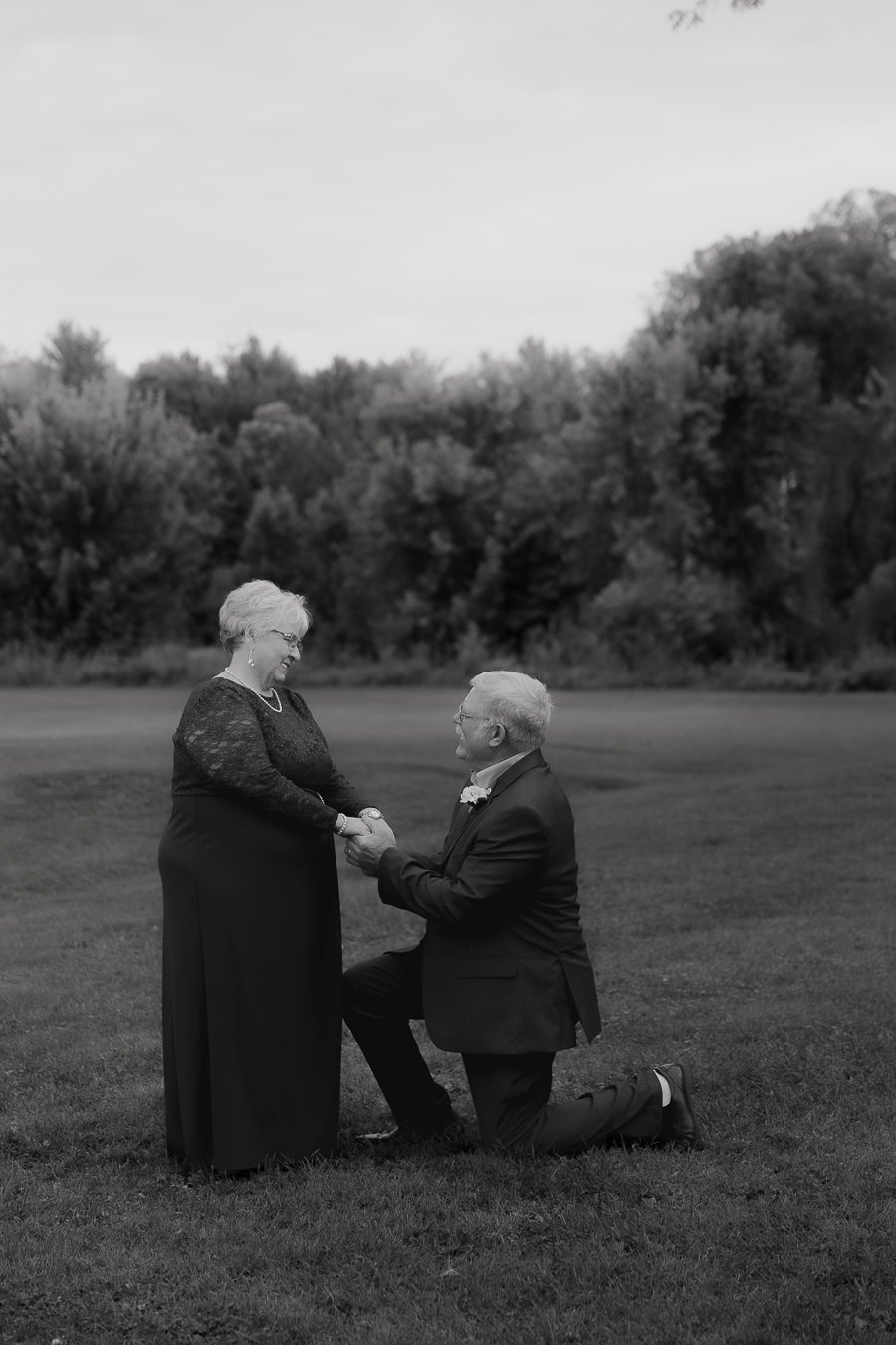 A man in a suit kneeling on one knee, holding hands with a woman in a long dress, outdoors on a grassy field with trees in the background.
