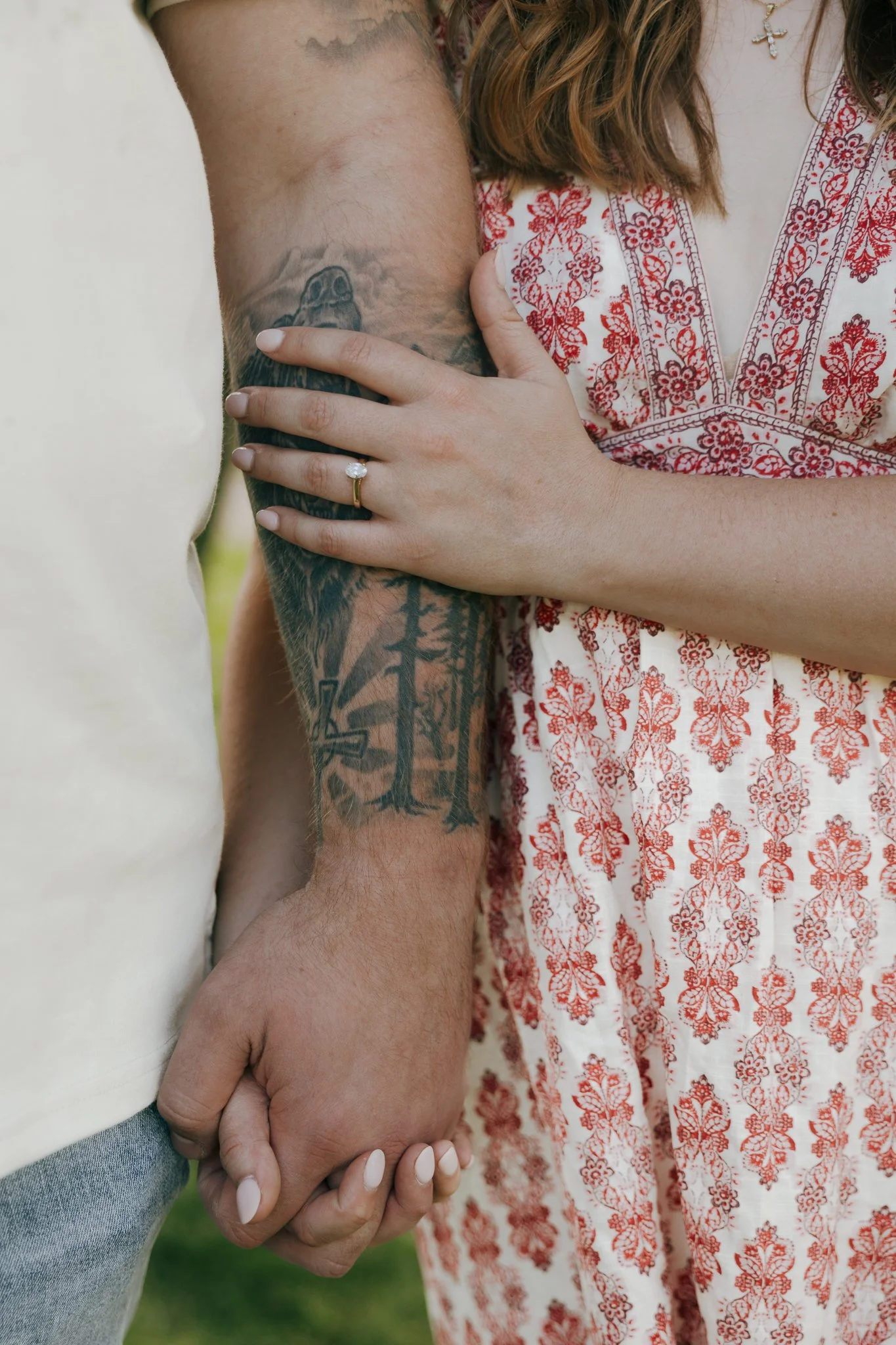 A couple holding hands, with the woman's hand resting on the man's arm. She is wearing a ring and has her nails painted white. The man has a tattoo on his forearm, and the woman is wearing a white dress with red floral patterns.