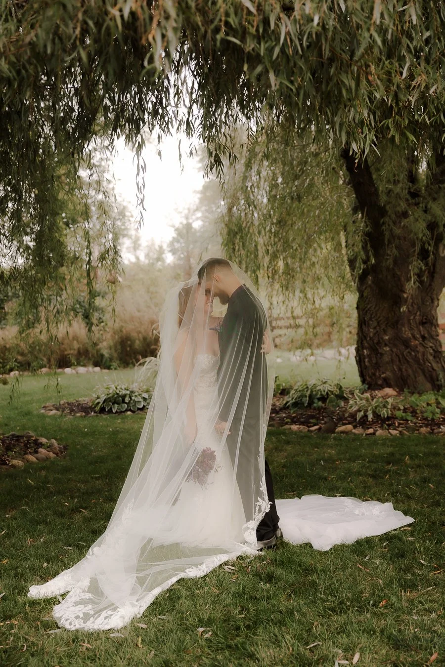 A bride and groom sharing a kiss under a large tree on a grassy lawn, with the bride wearing a white wedding gown and veil, holding a bouquet, and the groom in a dark suit with pinstripes, during their wedding.
