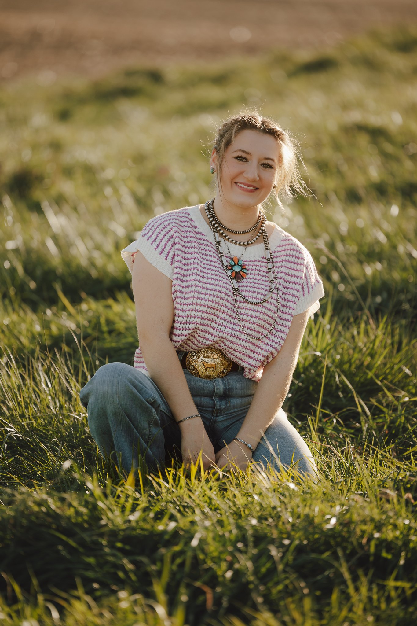 A smiling woman with blonde hair in a braid, wearing a striped pink and white top, and layered necklaces, sits outdoors on grass with a blurred green field background during golden hour.