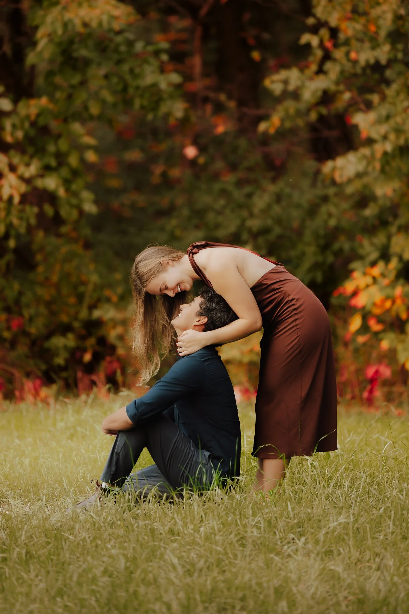 A couple sharing a joyful moment outdoors, with the woman bending down smiling at the man who is sitting on the grass, surrounded by green and autumn-colored foliage.