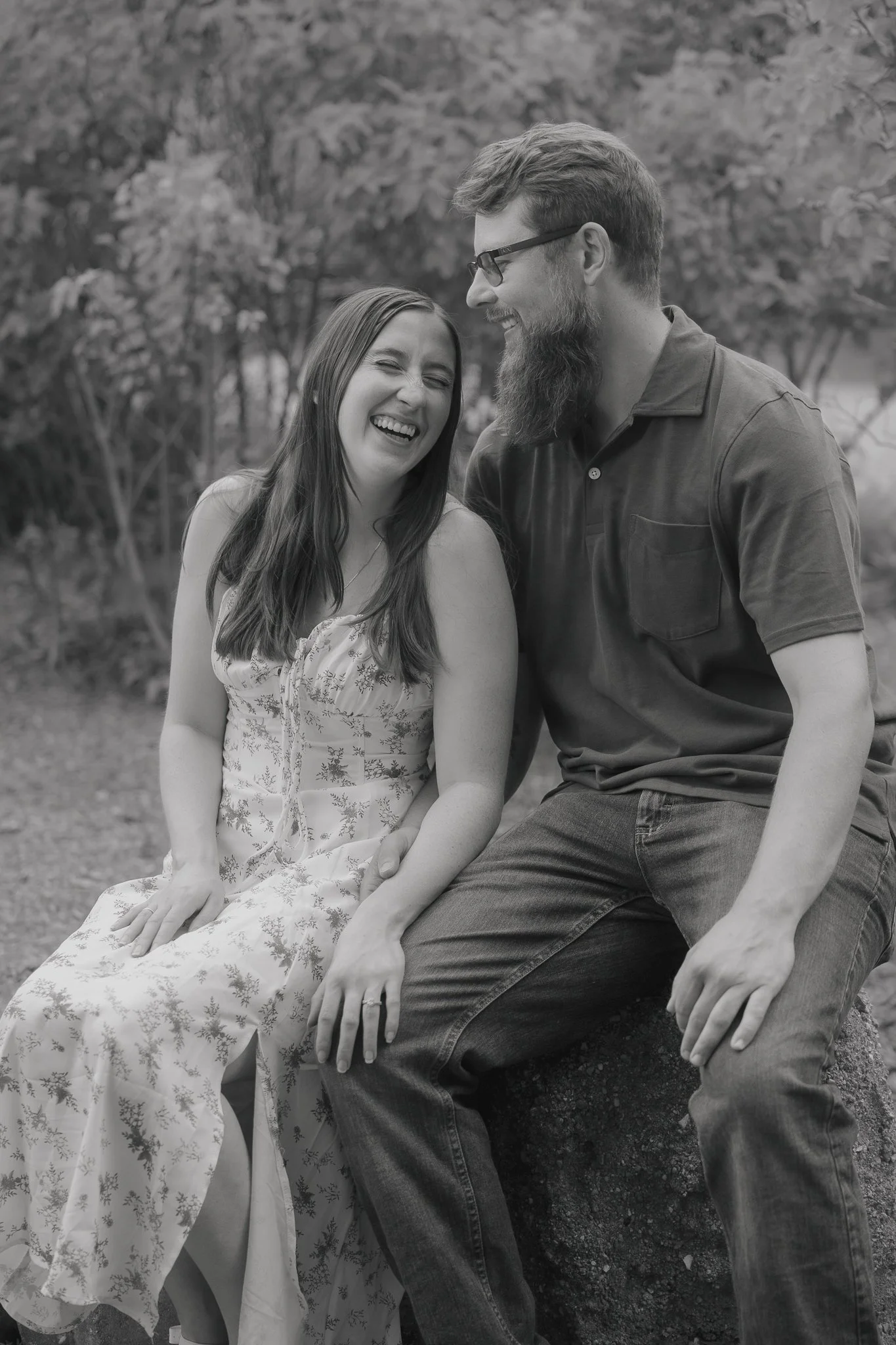 A black and white photo of a man and woman sitting outdoors, laughing together. The woman is wearing a patterned dress, and the man has a beard and glasses, dressed in a polo shirt and jeans.