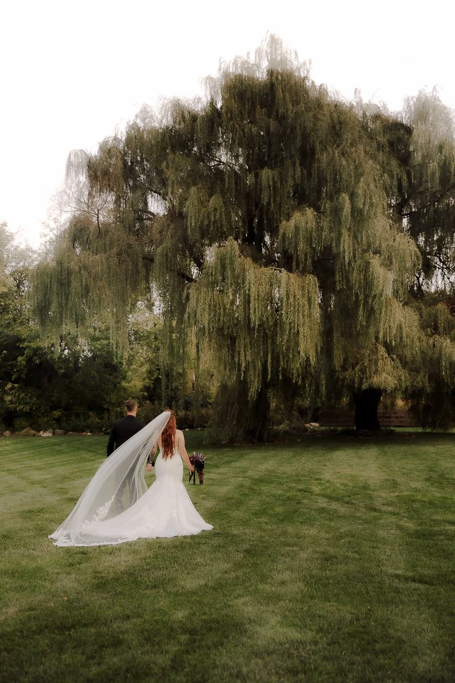 A bride and groom holding hands and walking on a grassy field, with the bride in a white wedding gown and veil, carrying a bouquet, and large trees in the background.