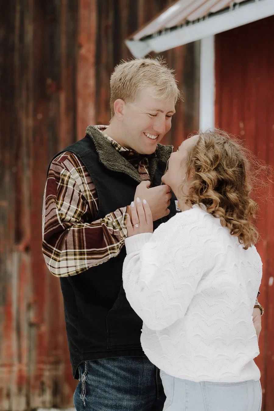 A happy couple smiling at each other outdoors in front of a red barn, with the man touching the woman's chin gently.