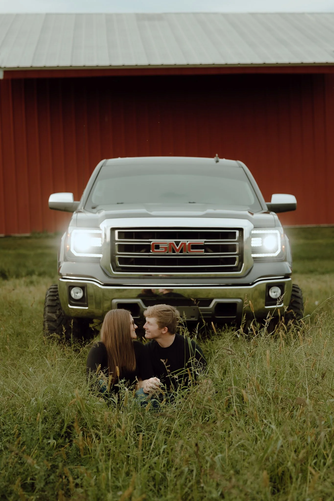 A silver GMC truck parked on grass in front of a red barn, with a couple sitting in the grass in front of the truck.