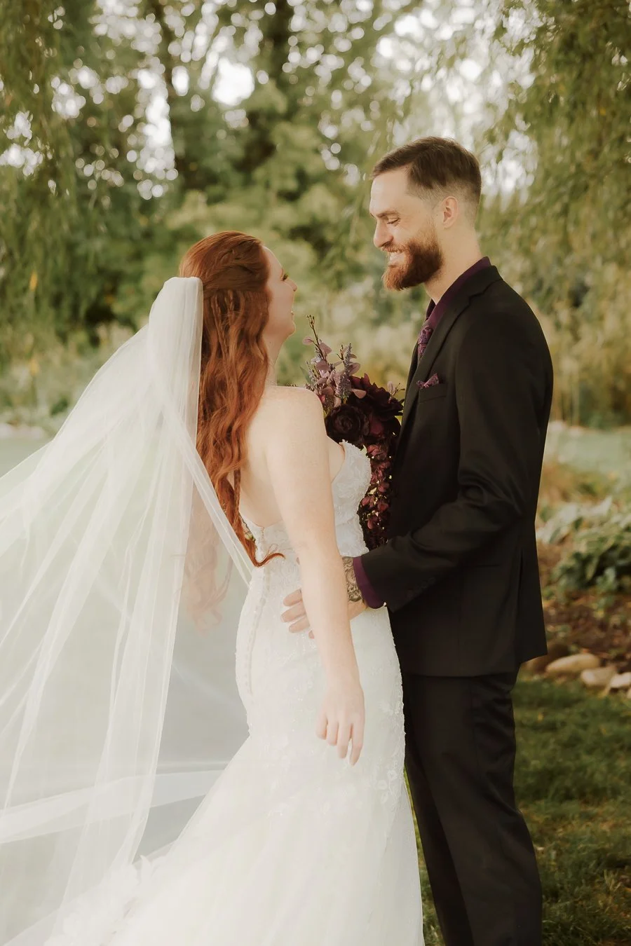 A bride with long red hair, wearing a white wedding gown and veil, facing a groom with short dark hair, beard, and wearing a black suit, standing outdoors with trees in the background.