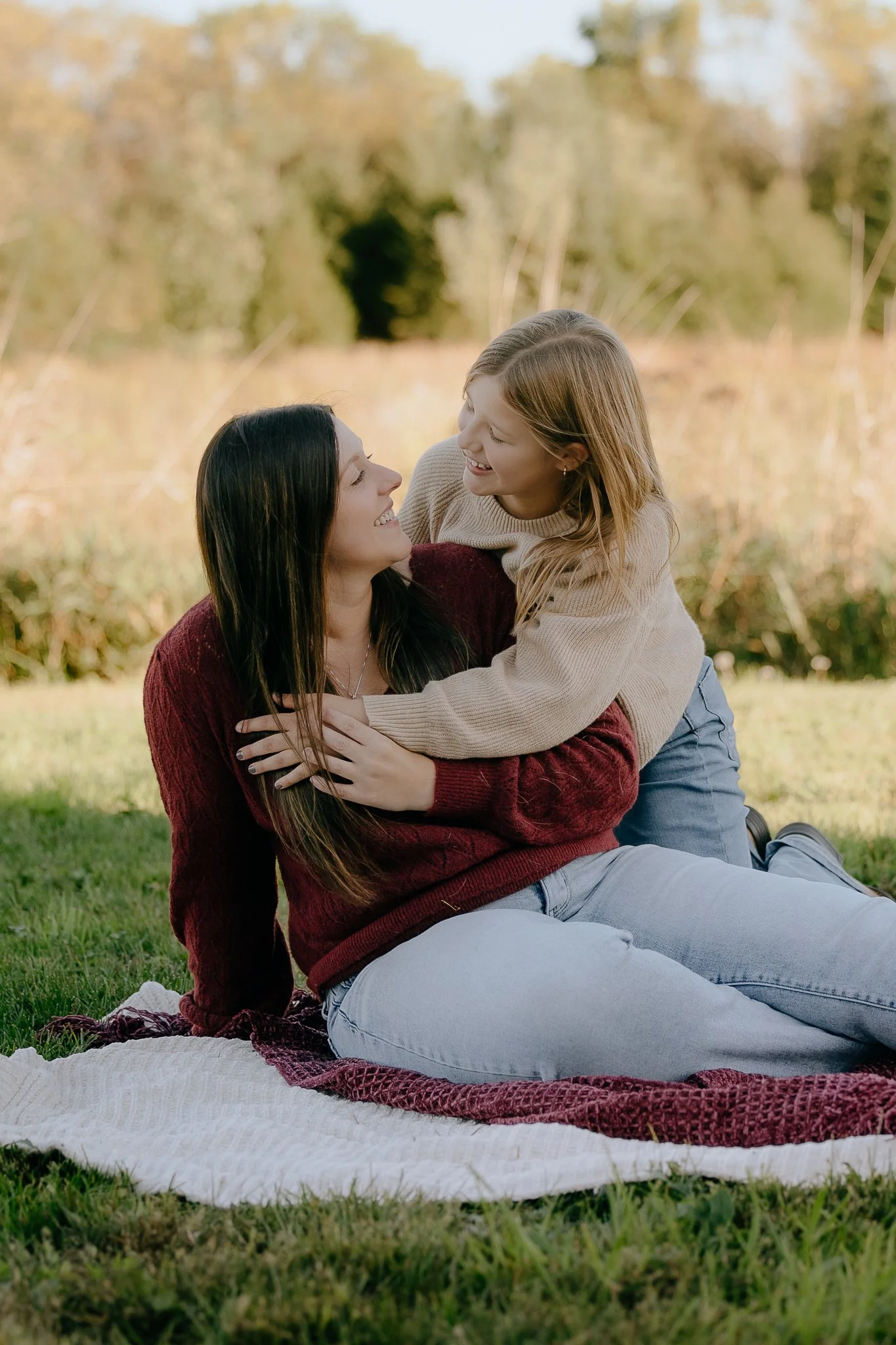 A woman and a young girl are sitting on a blanket outdoors, smiling and embracing each other in a grassy area with trees in the background.
