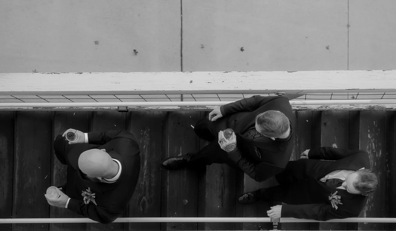 Three men in formal suits holding drinks, standing and talking on a black wooden floor in an industrial or warehouse-like setting.