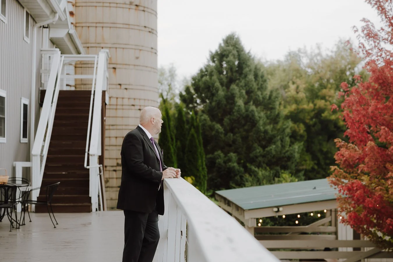 A man in a black suit and purple tie stands on a balcony, looking out at lush green trees with some red leaves, during daytime.