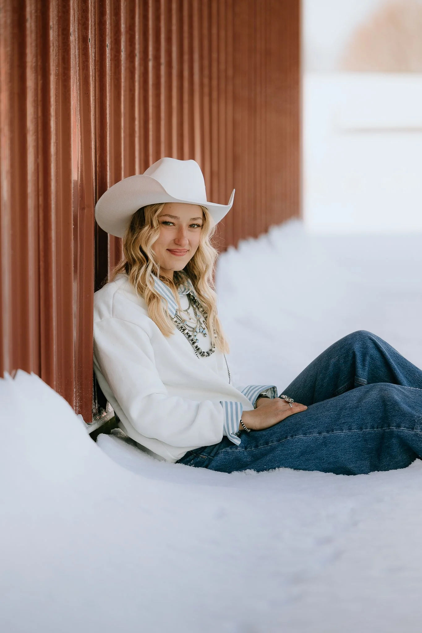 A young woman sitting in the snow, leaning against a red wooden wall, wearing a white cowboy hat, white sweater, striped shirt, jeans, and jewelry, smiling at the camera.