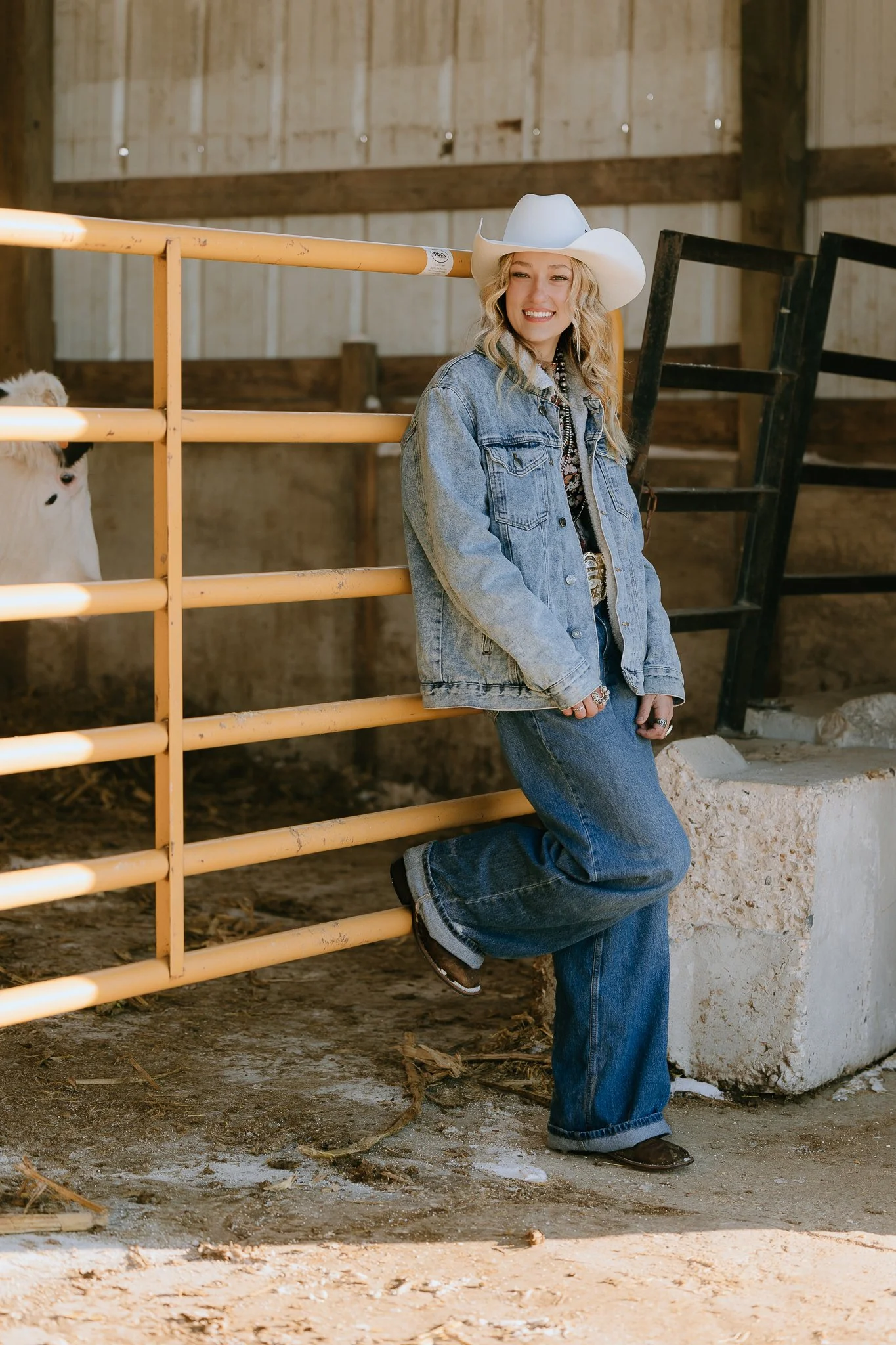 A woman wearing a white cowboy hat, denim jacket, and jeans, smiling while standing by a yellow livestock fence inside a barn, with a calf visible behind the fence.