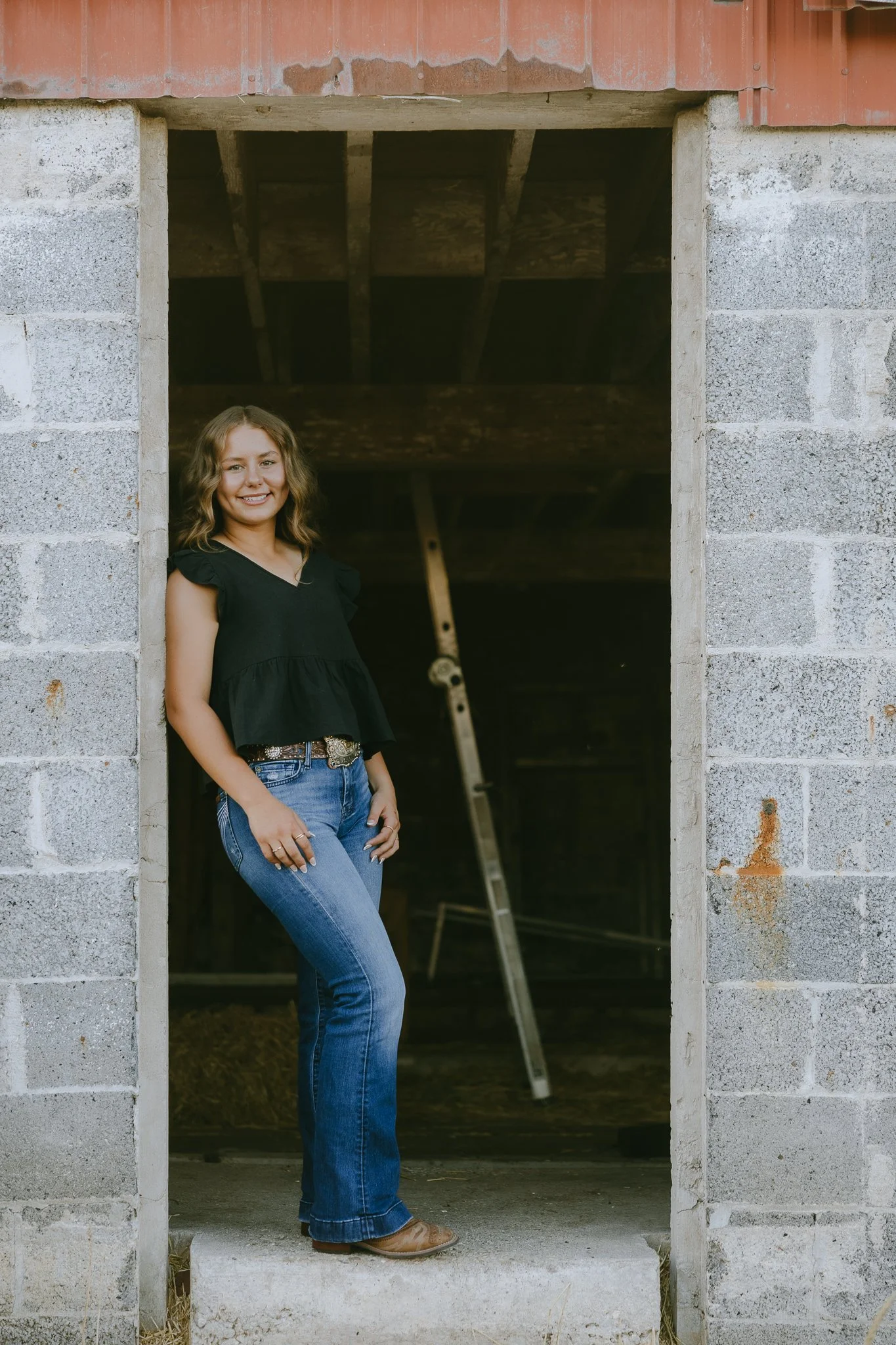 Young woman standing at the entrance of a building under construction, smiling, wearing a black top and blue jeans.