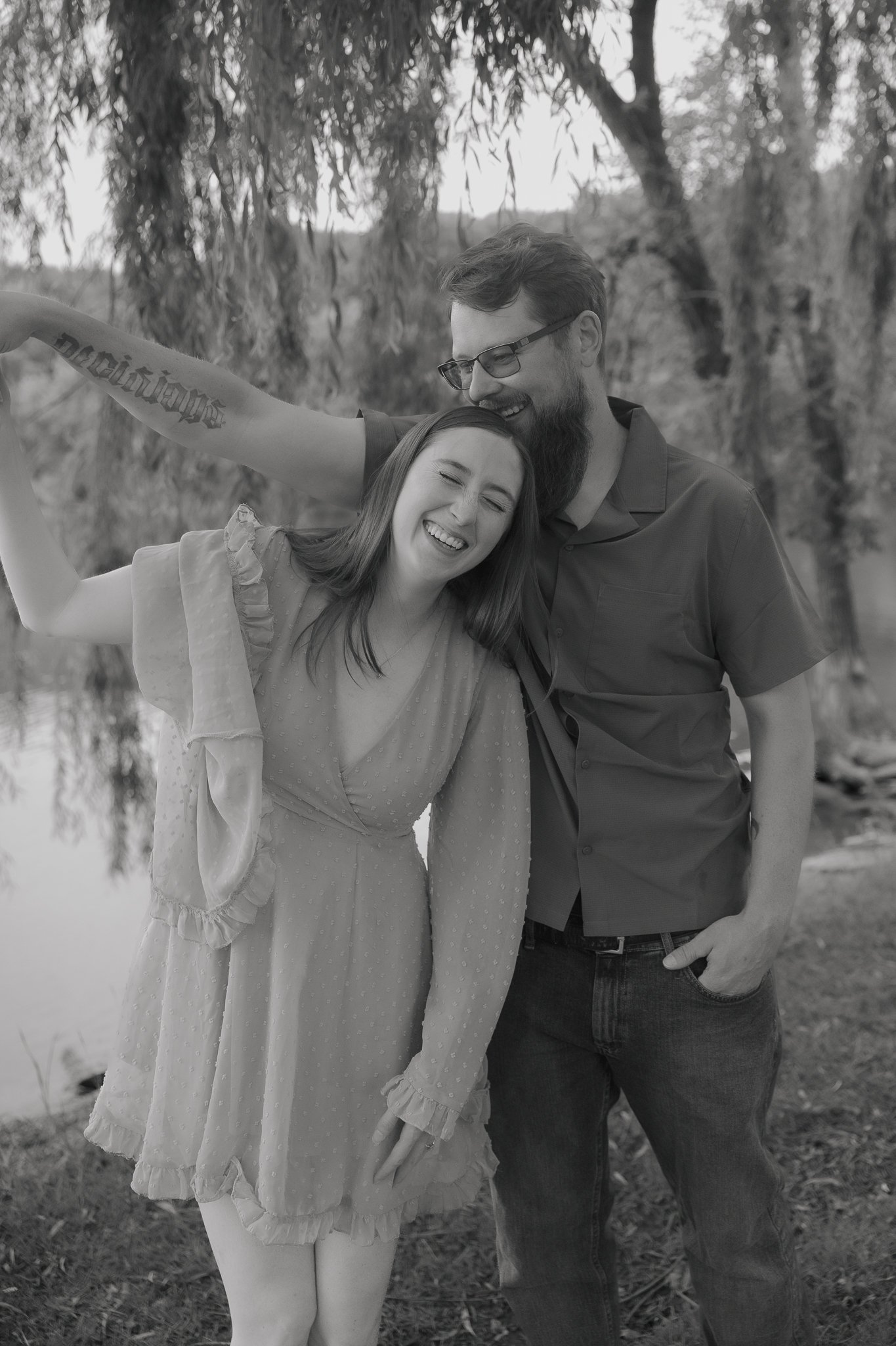A black and white photo of a smiling couple outdoors near a body of water, with trees in the background. The woman has shoulder-length hair and is wearing a light dress with ruffled sleeves. The man has a beard, glasses, and is wearing a dark short-s