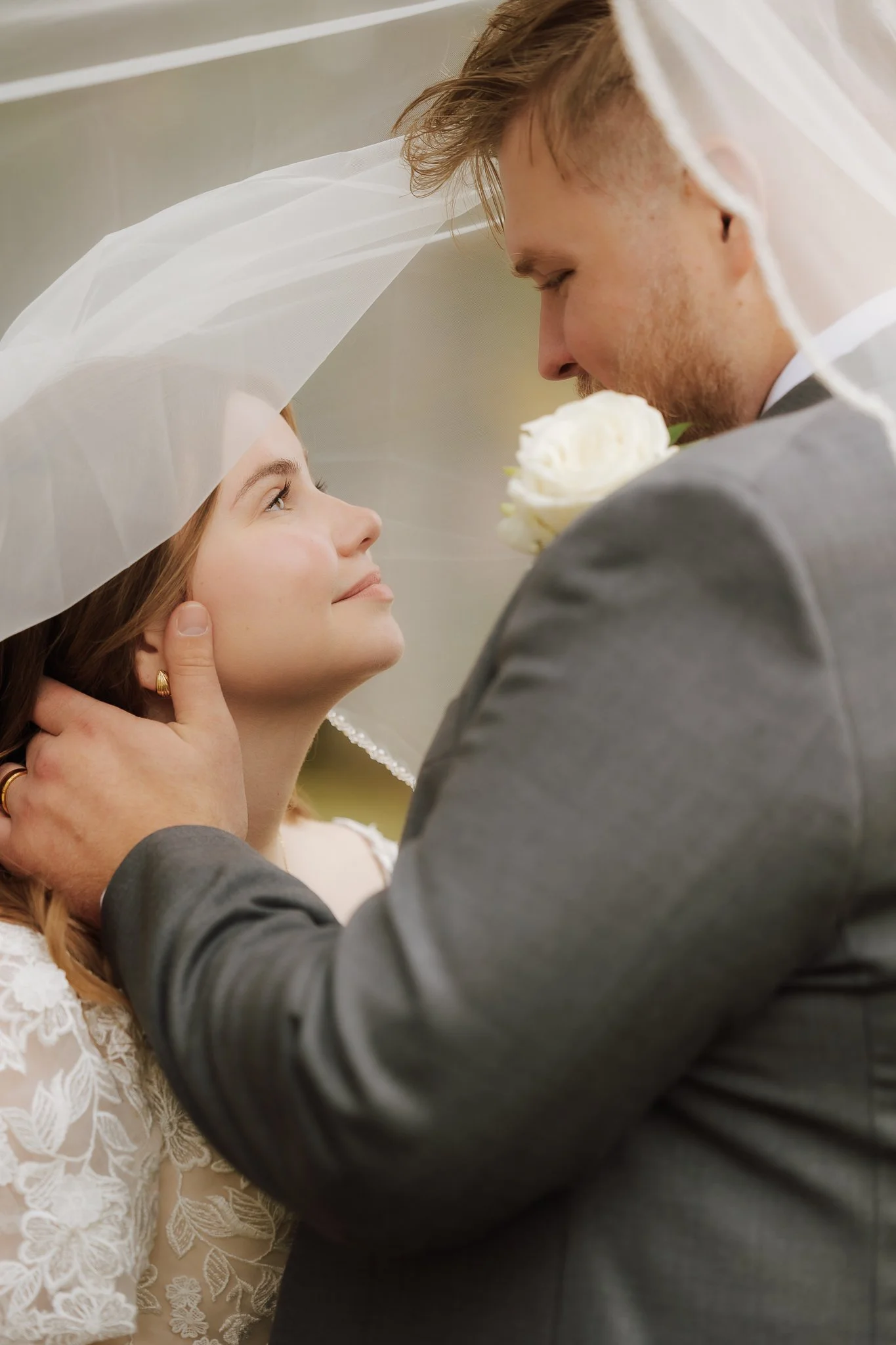 A bride and groom sharing an intimate moment, with the groom holding the bride's face and looking into her eyes. The bride wears a veil and a white lace dress, and the groom wears a gray suit and holds a white rose.