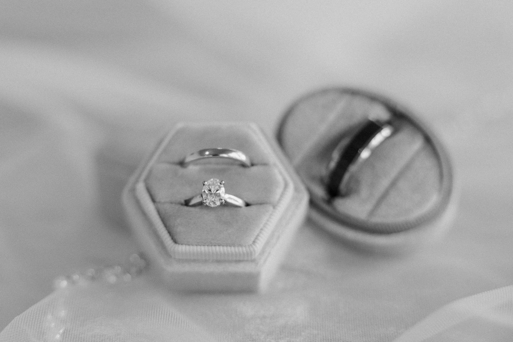 Engagement ring with a large diamond in a velvet ring box, with a wedding band on top of a ring box and another wedding band inside an open ring box. All rings are metallic, possibly silver or platinum, on a soft fabric surface.