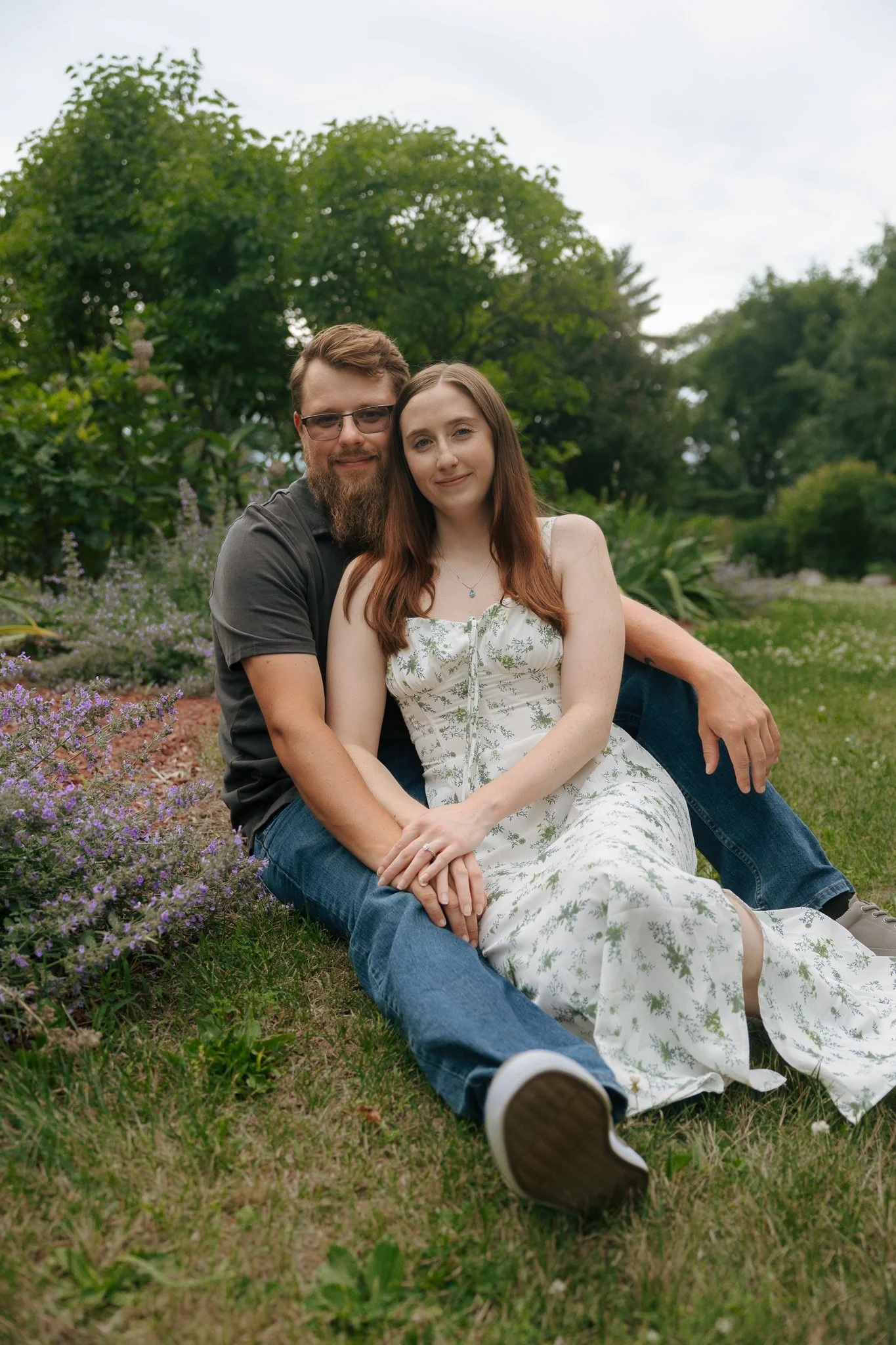 A young couple sitting on the grass in a park, surrounded by greenery and purple flowers, with a large tree behind them, enjoying a peaceful outdoor moment.