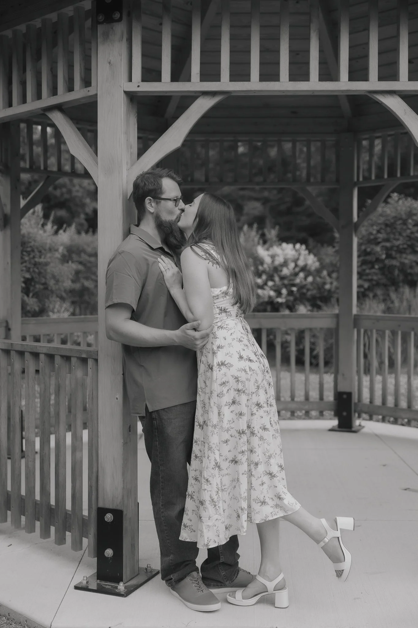 A black and white photo of a couple kissing under a wooden gazebo. The woman is wearing a floral dress and high heels, while the man is in a casual shirt and jeans.