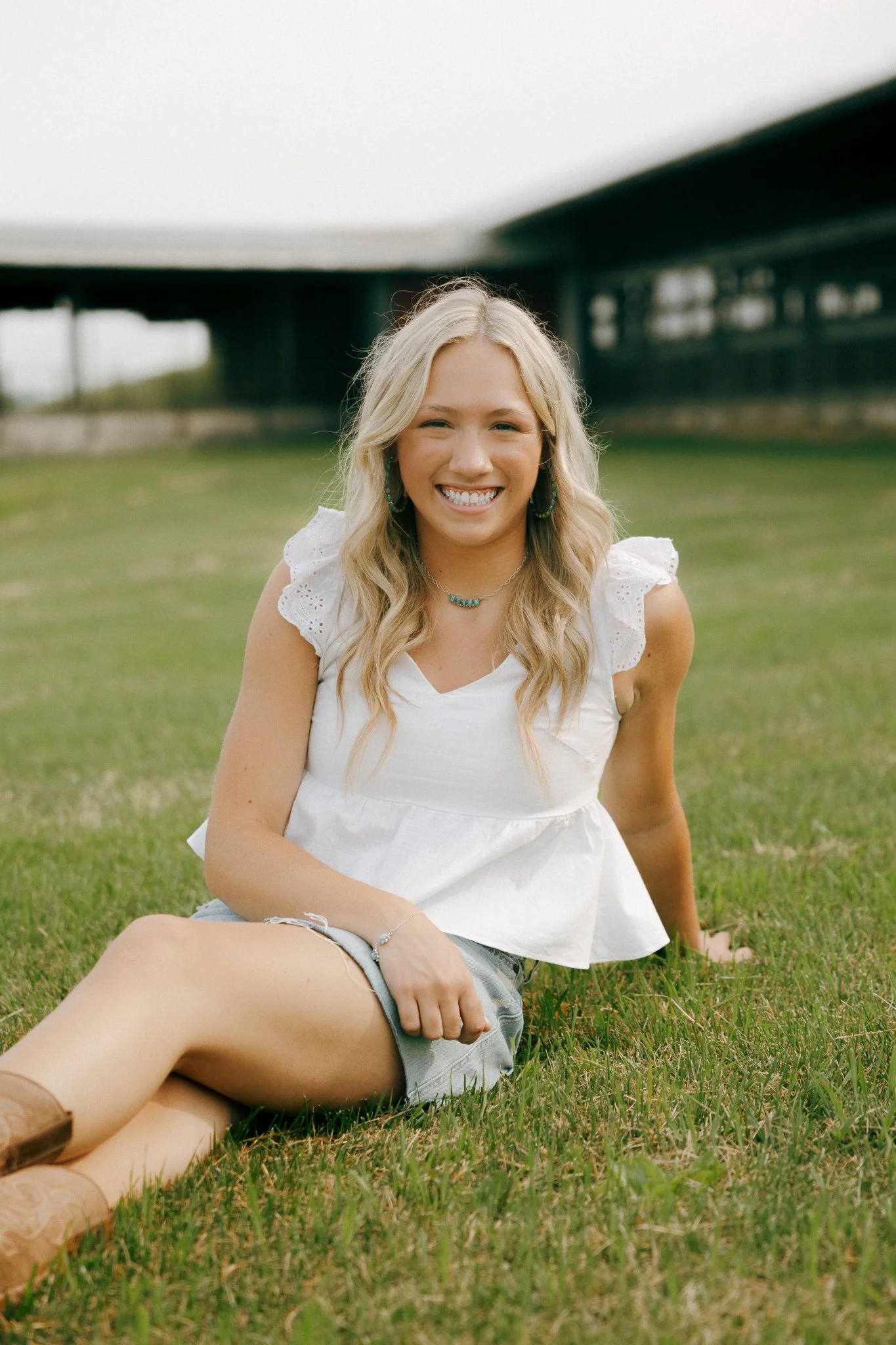 A young woman with long blonde hair smiling and sitting on the grass outdoors under a bridge.