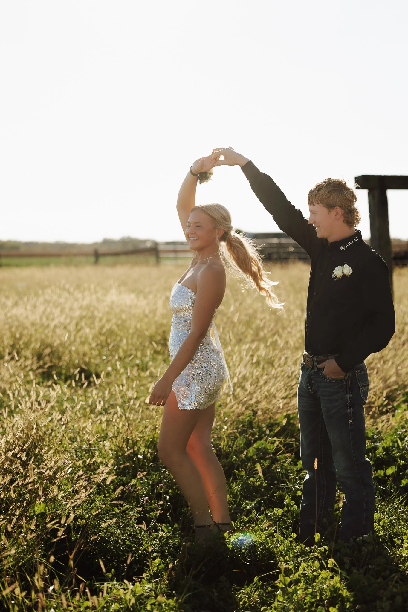 A couple is dancing in a grassy field during sunset, with the girl in a shiny dress and the boy in a black shirt. The boy twirls the girl by holding her hand up as she smiles, surrounded by tall grass and farm fencing in the background.