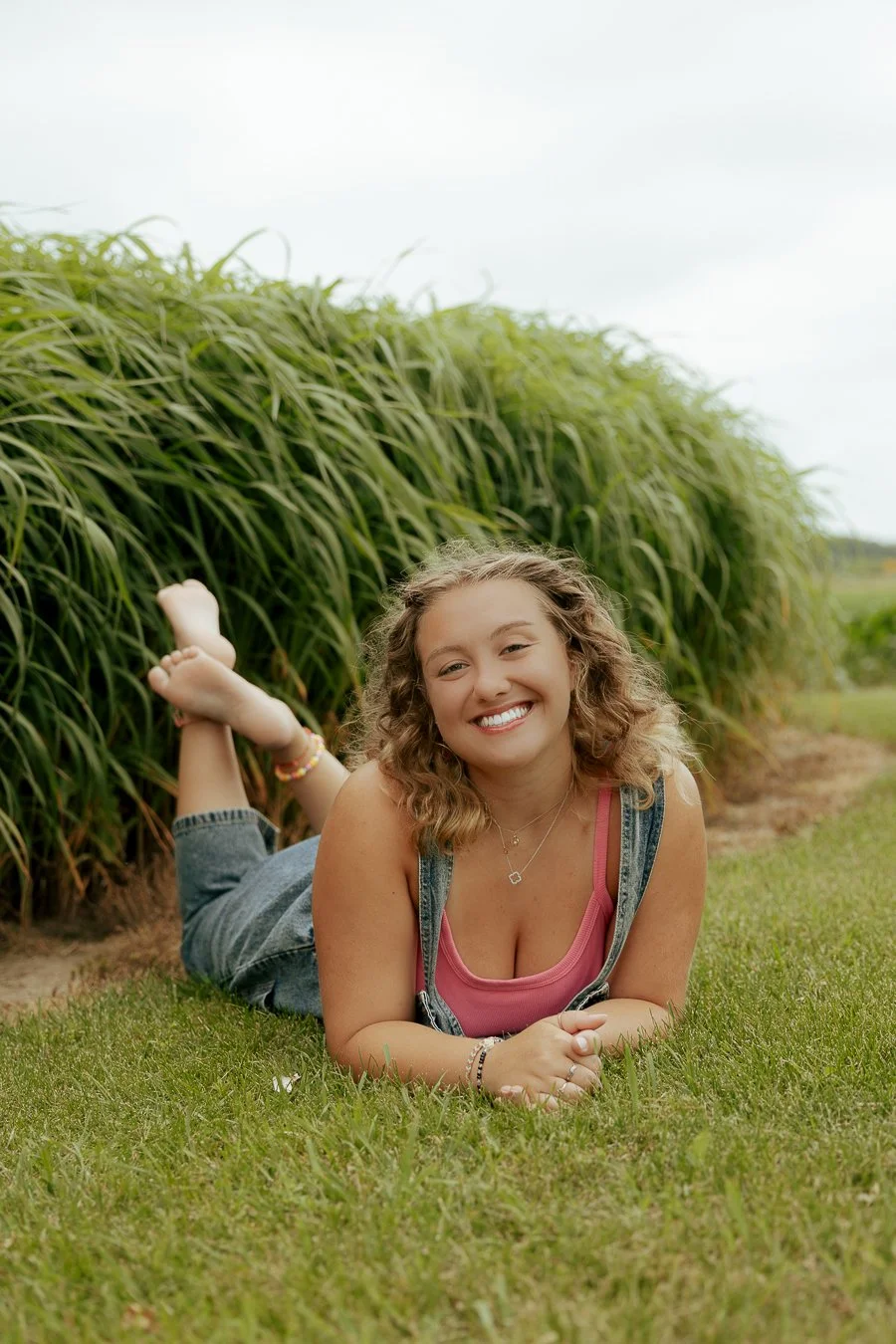 A young woman with curly hair lying on her stomach on a grassy field, smiling, with tall green plants in the background under an overcast sky.