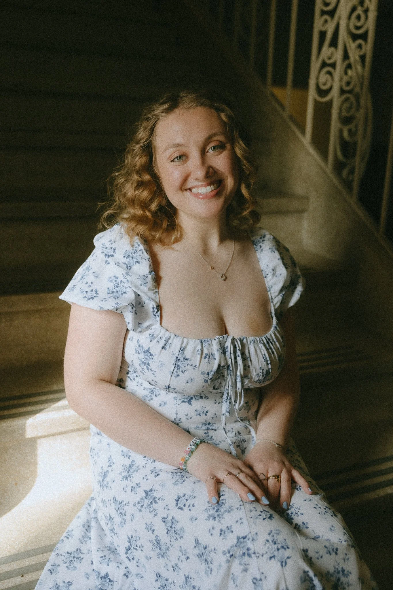 A young woman with curly red hair and fair skin, smiling, seated on a staircase with a decorative iron railing, wearing a white dress with blue floral pattern and jewelry.