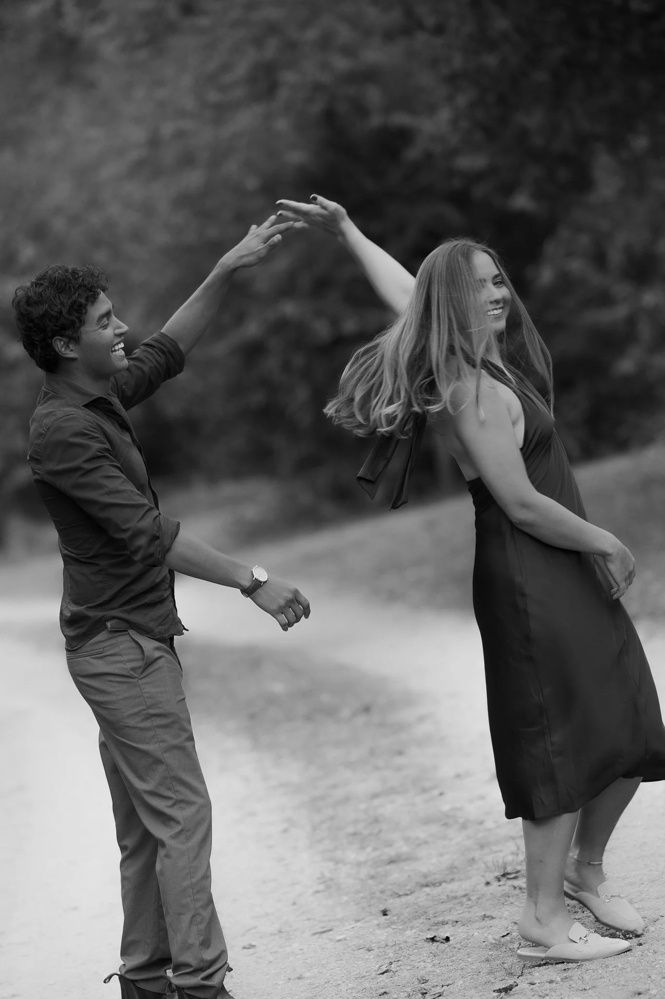 A black and white photo of a young man and woman dancing outdoors. The man is smiling at the woman, reaching out his hand, while the woman is spinning or twirling, smiling, with her hair flowing. The background shows trees and a dirt path.