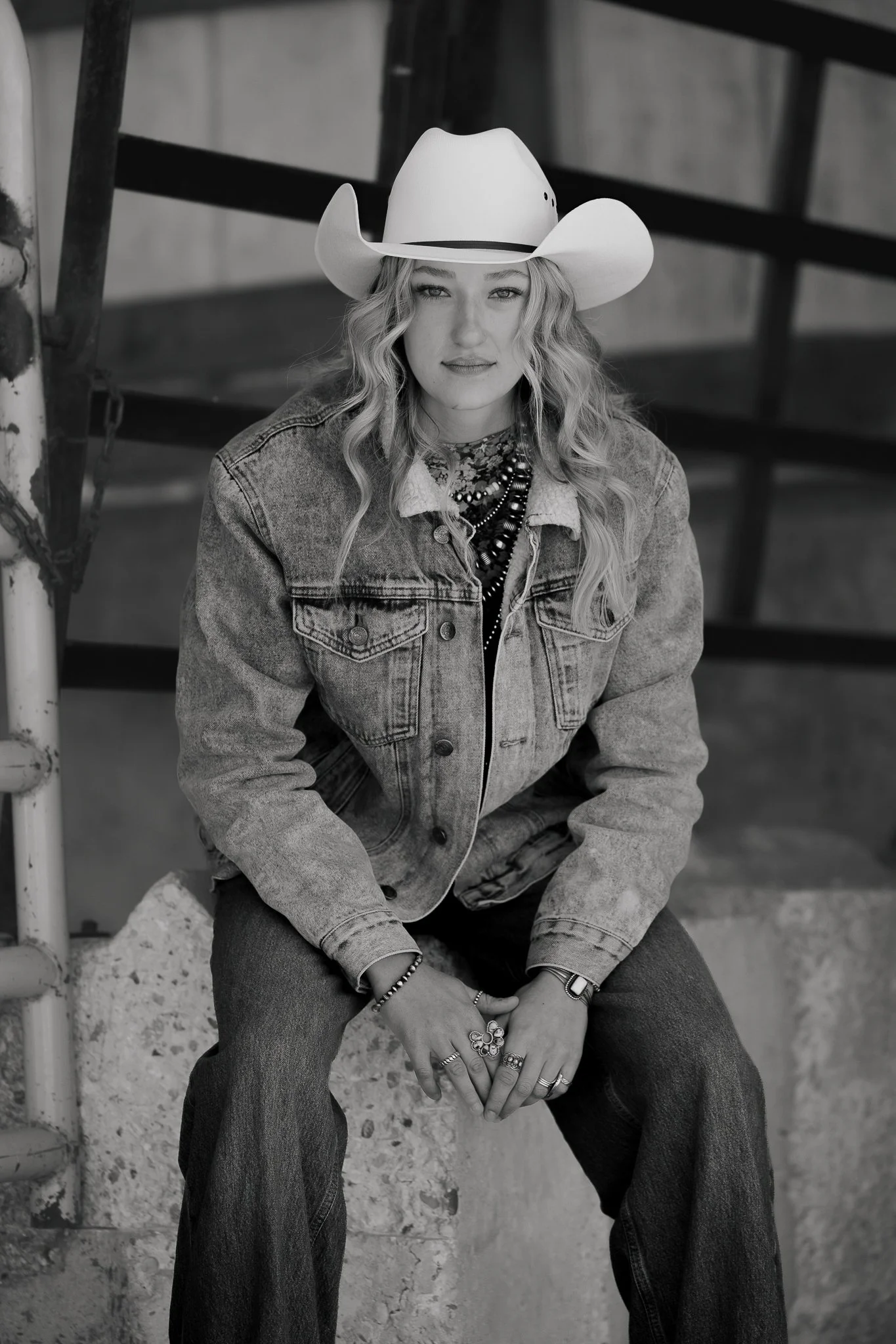 Black and white photo of a young woman with long wavy hair, wearing a cowboy hat, denim jacket, and multiple rings and necklaces, sitting on a concrete block with a metal staircase behind her.