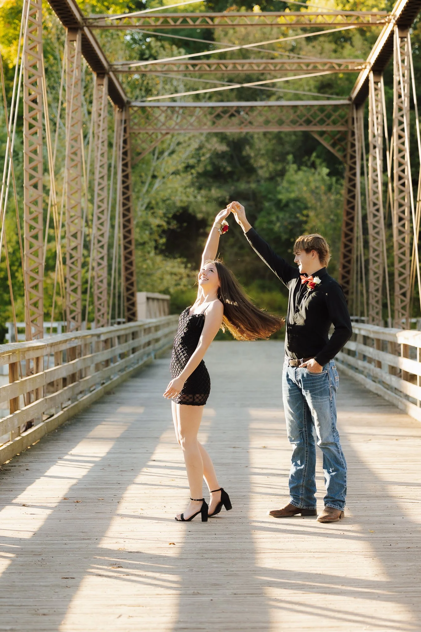 A couple dancing on a bridge outdoors during daytime, with the man twirling the woman in a black dress and heels, surrounded by green trees and sunlight.