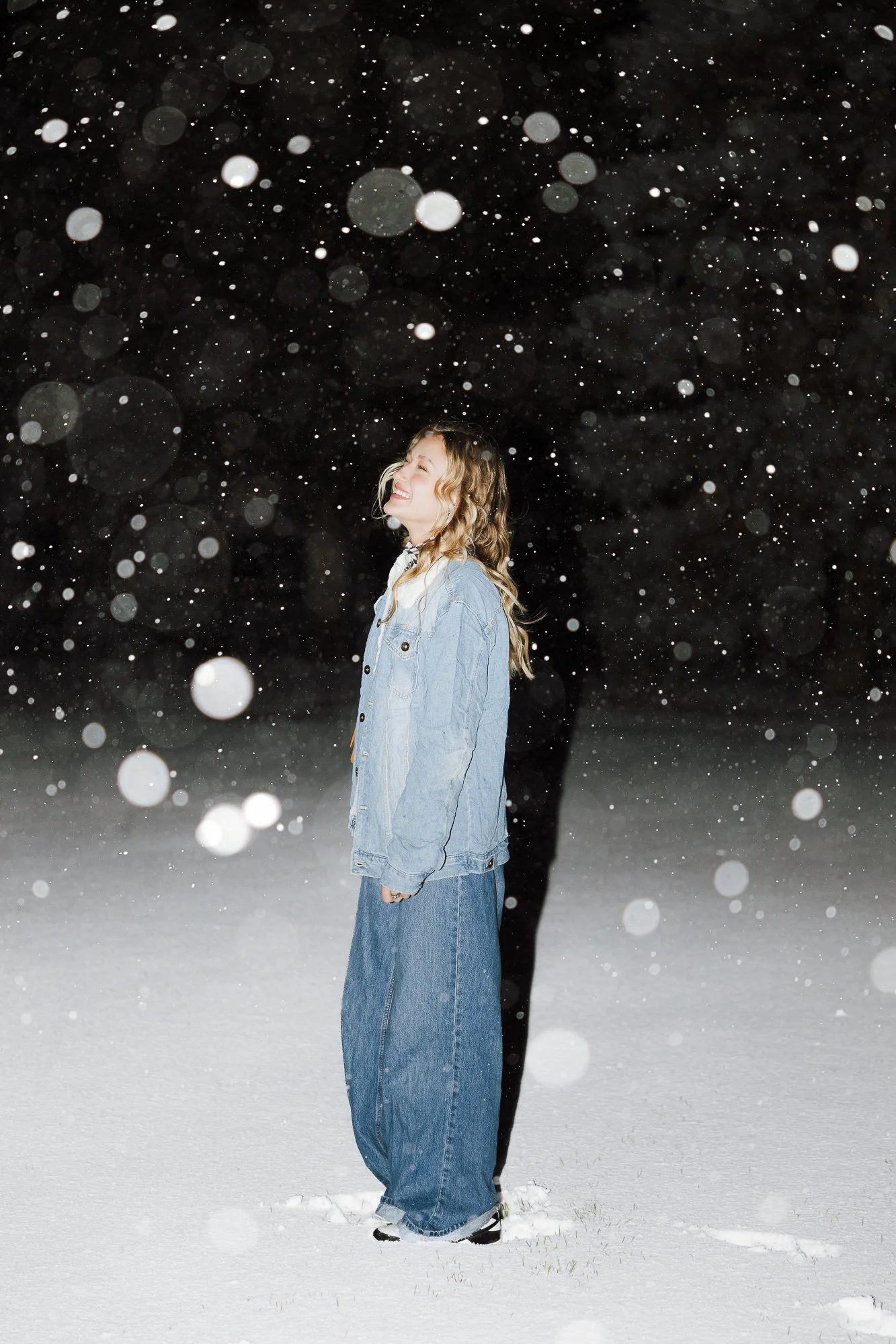 A woman smiling and standing in a snowy field at night with snow falling around her.