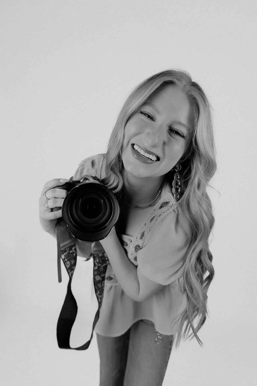 A smiling woman with long, wavy hair taking a selfie with a camera against a plain background.