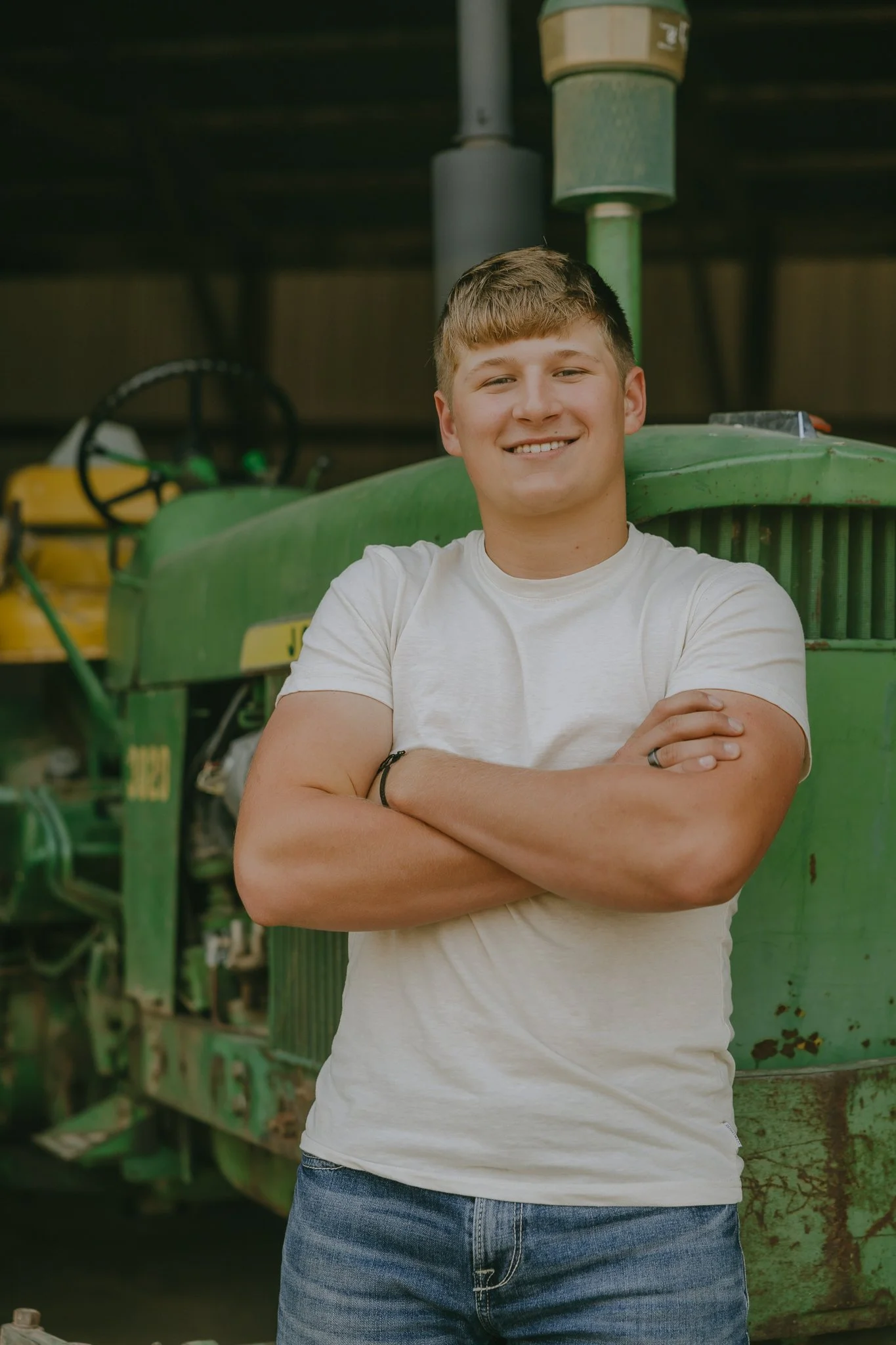 A young man with short blonde hair, wearing a white t-shirt and jeans, standing with arms crossed in front of a green tractor, smiling at the camera inside a barn.