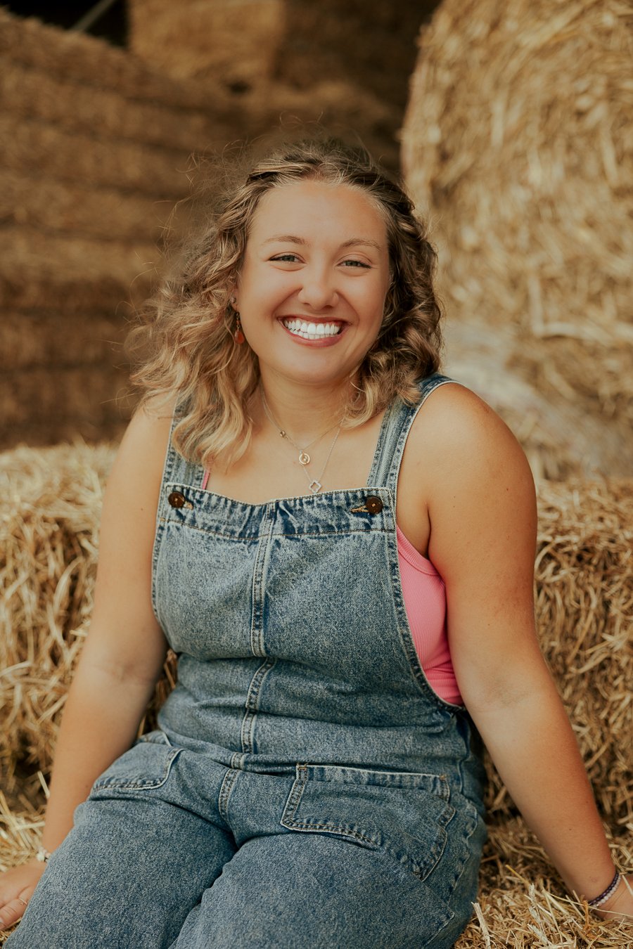 A woman with curly blonde hair and a bright smile, sitting among hay bales, wearing denim overalls over a pink shirt.