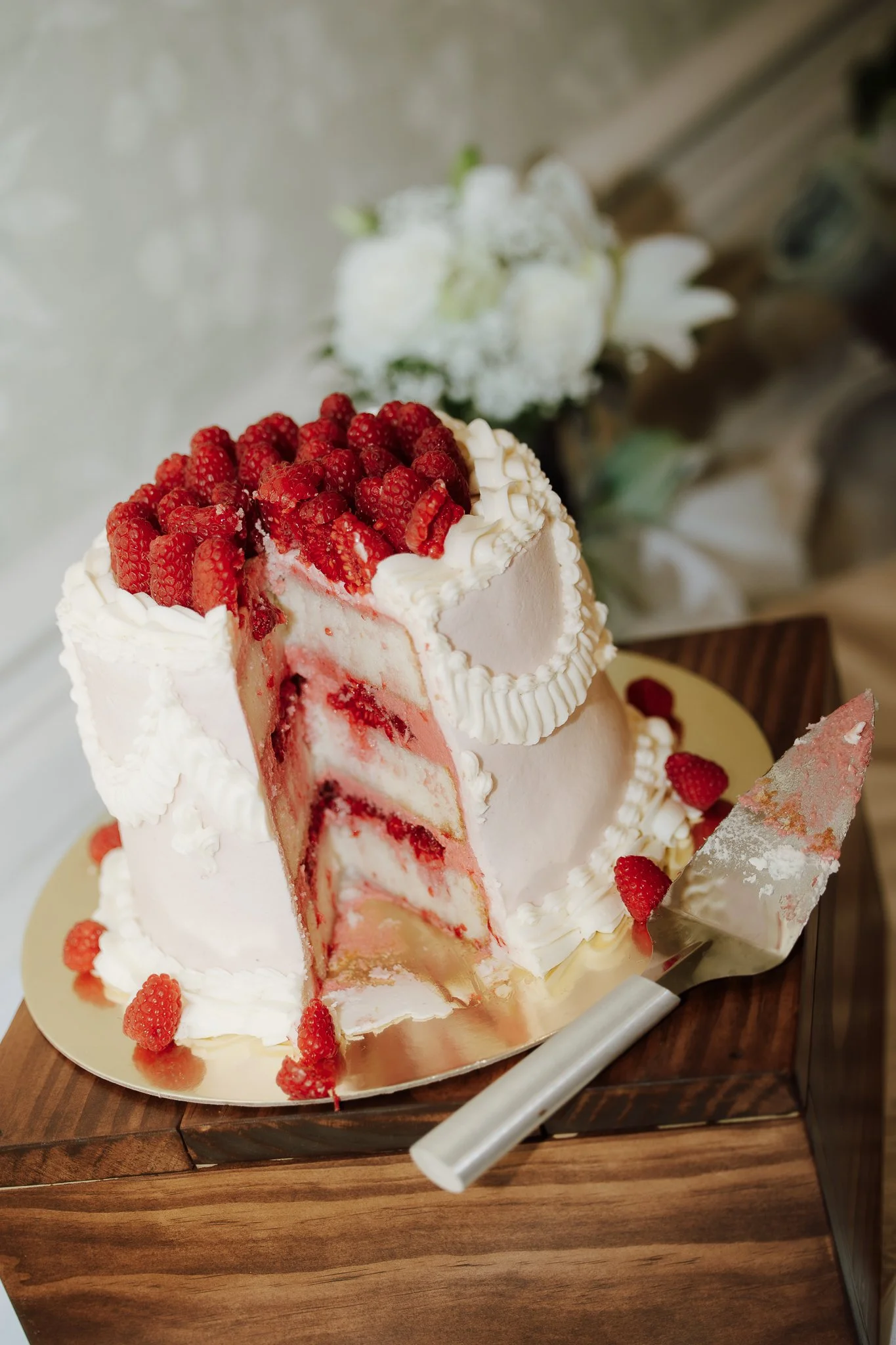 A partially sliced pink layered cake decorated with strawberries and small pieces of red fruit on top and around the base, sitting on a gold-colored cake board on a wooden surface with a cake server nearby.