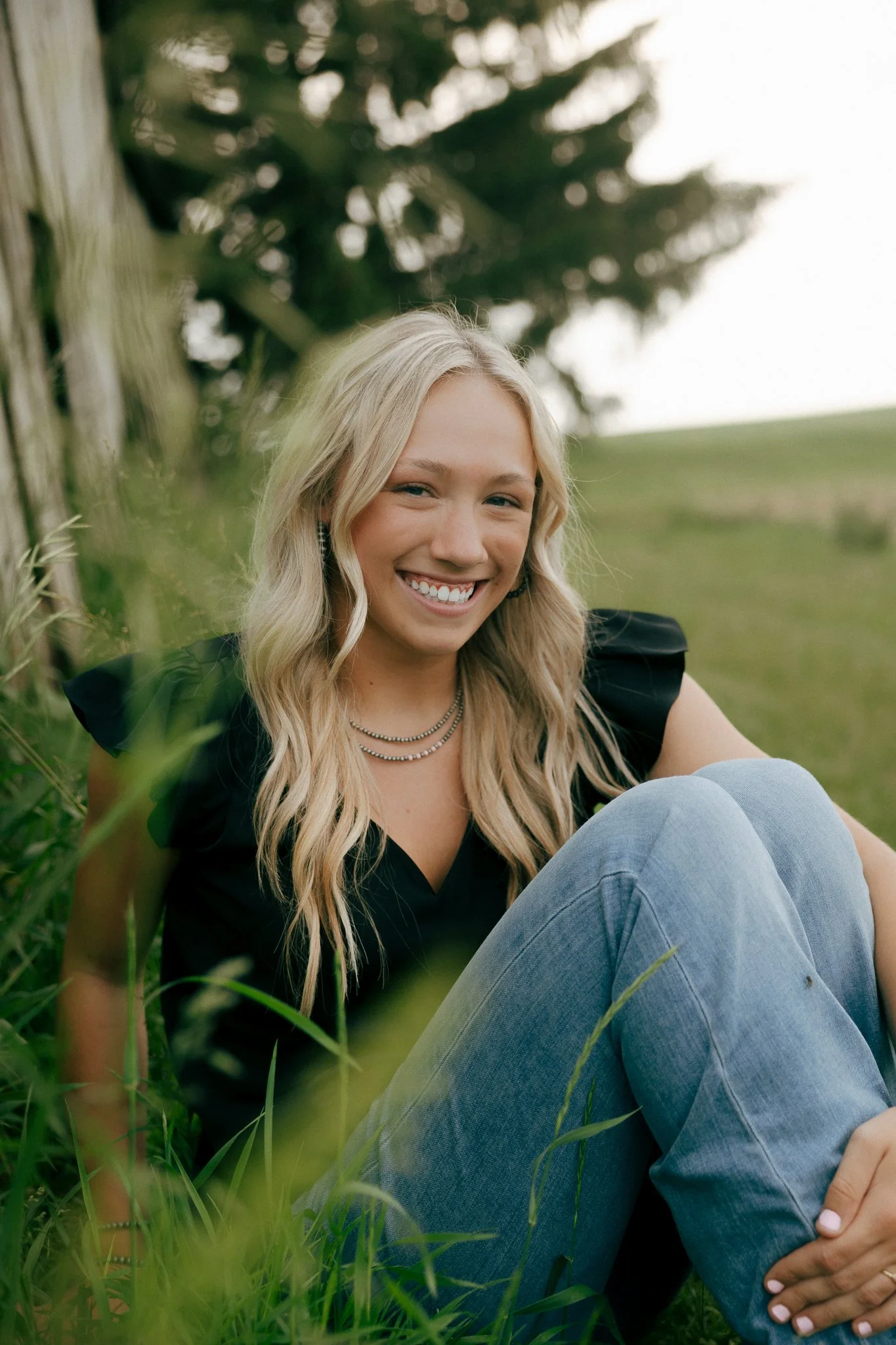 A young woman with long blonde hair, wearing a black top and light blue jeans, sitting outdoors in a grassy area, smiling at the camera.