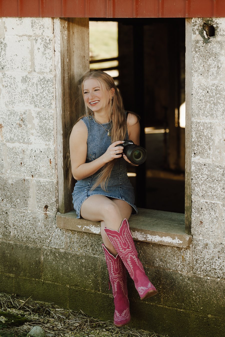 A young woman with long hair sitting on a window ledge of a rustic barn, wearing a denim dress, pink cowboy boots, and jewelry, holding a camera and smiling.