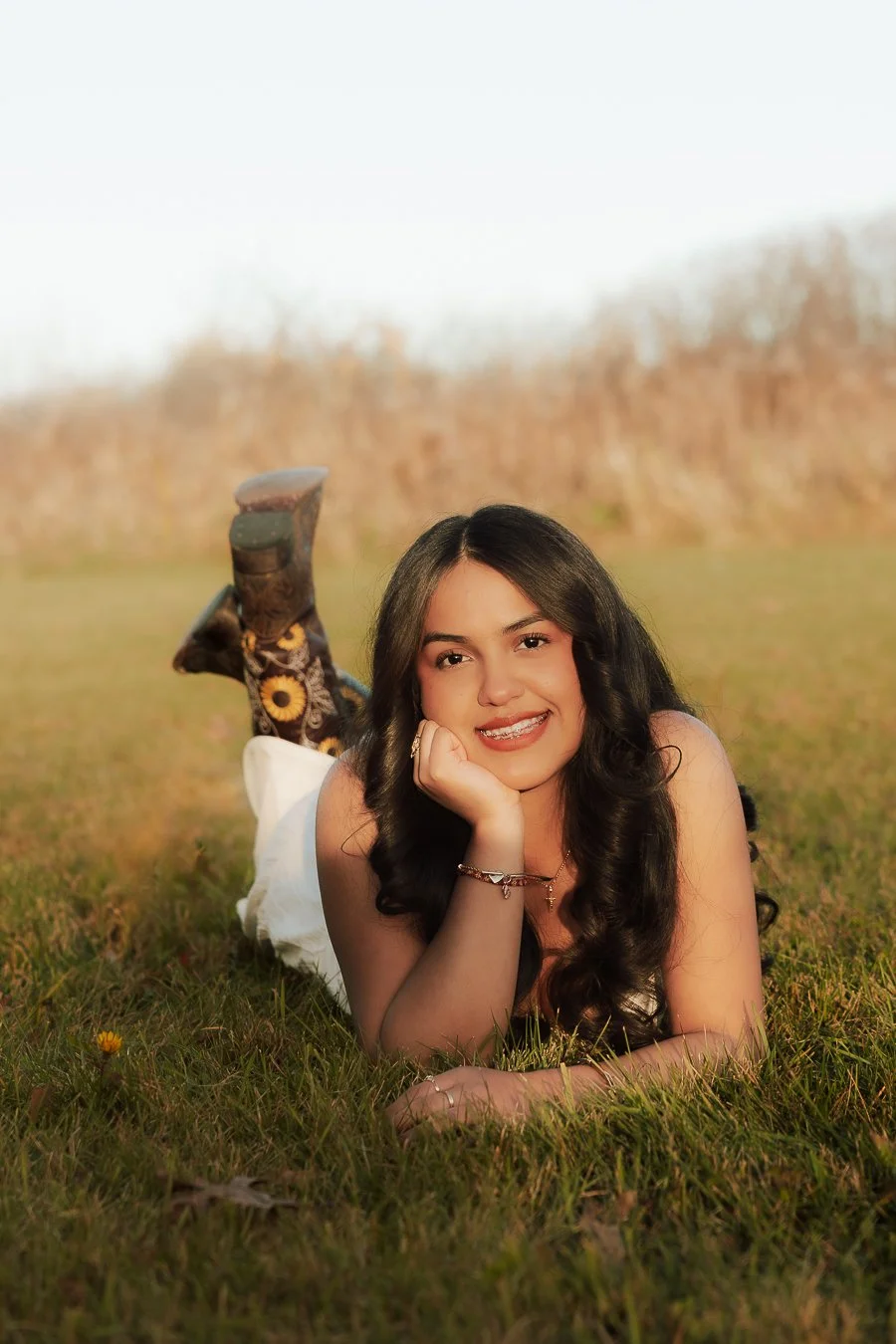 A young woman with long dark curly hair lying on her stomach on a grassy field, smiling at the camera, with a blurred natural background.