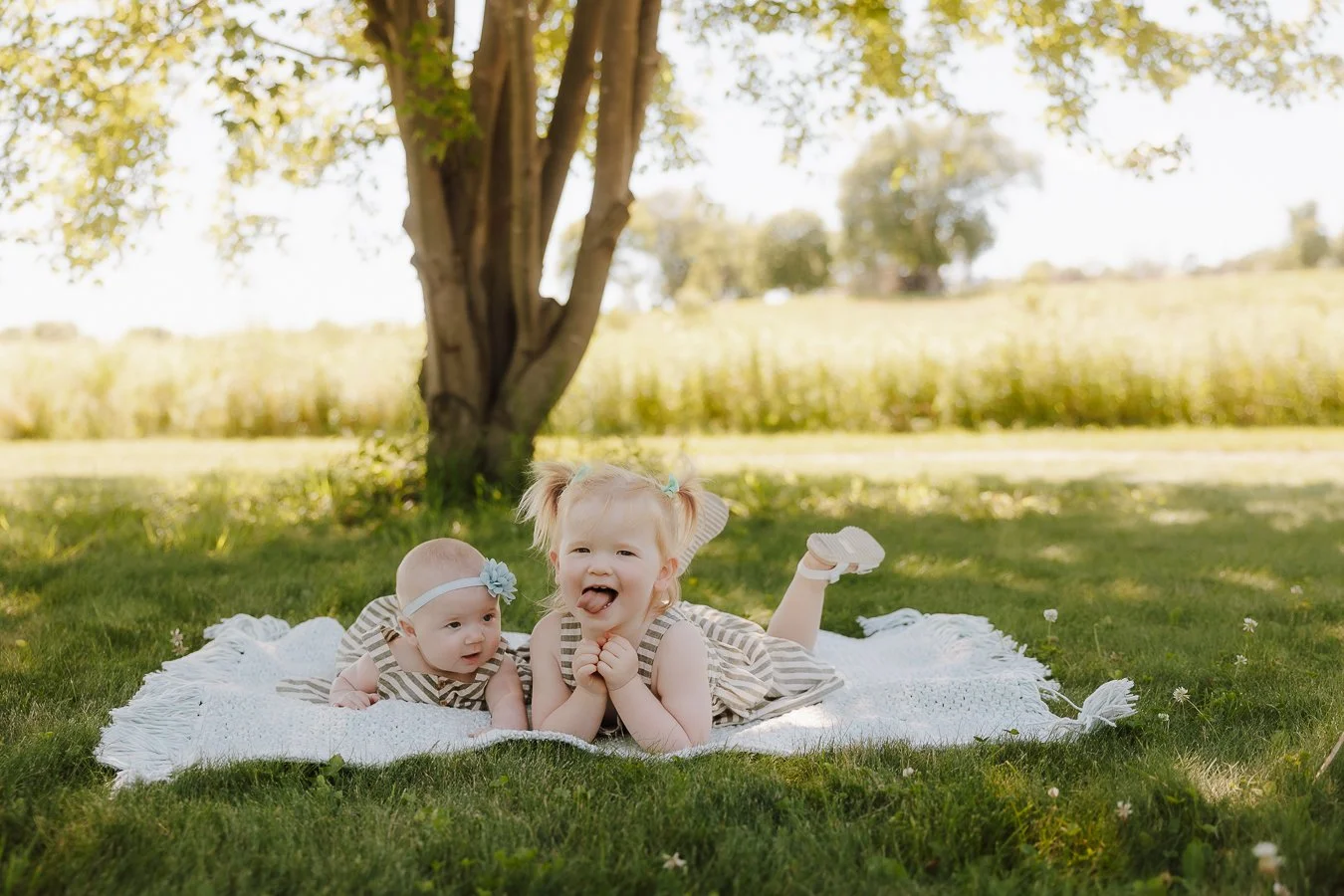 Two young children, a baby and a toddler girl, lying on a white blanket under a tree in a sunny park or field. The toddler has her tongue out and appears to be smiling, while the baby is looking forward. The scene is peaceful with grass and open land