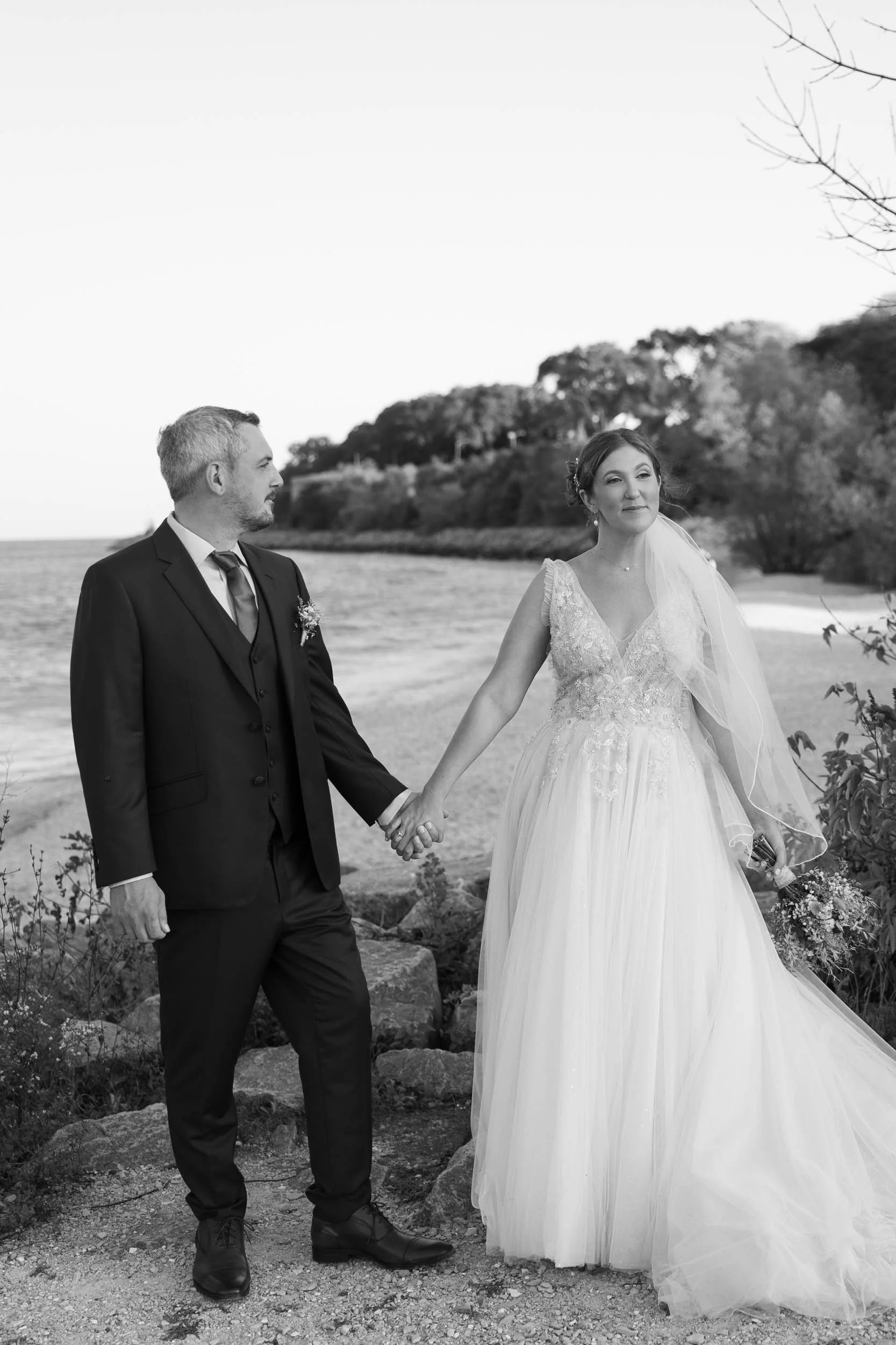A bride and groom holding hands by a body of water on a beach with trees in the background.