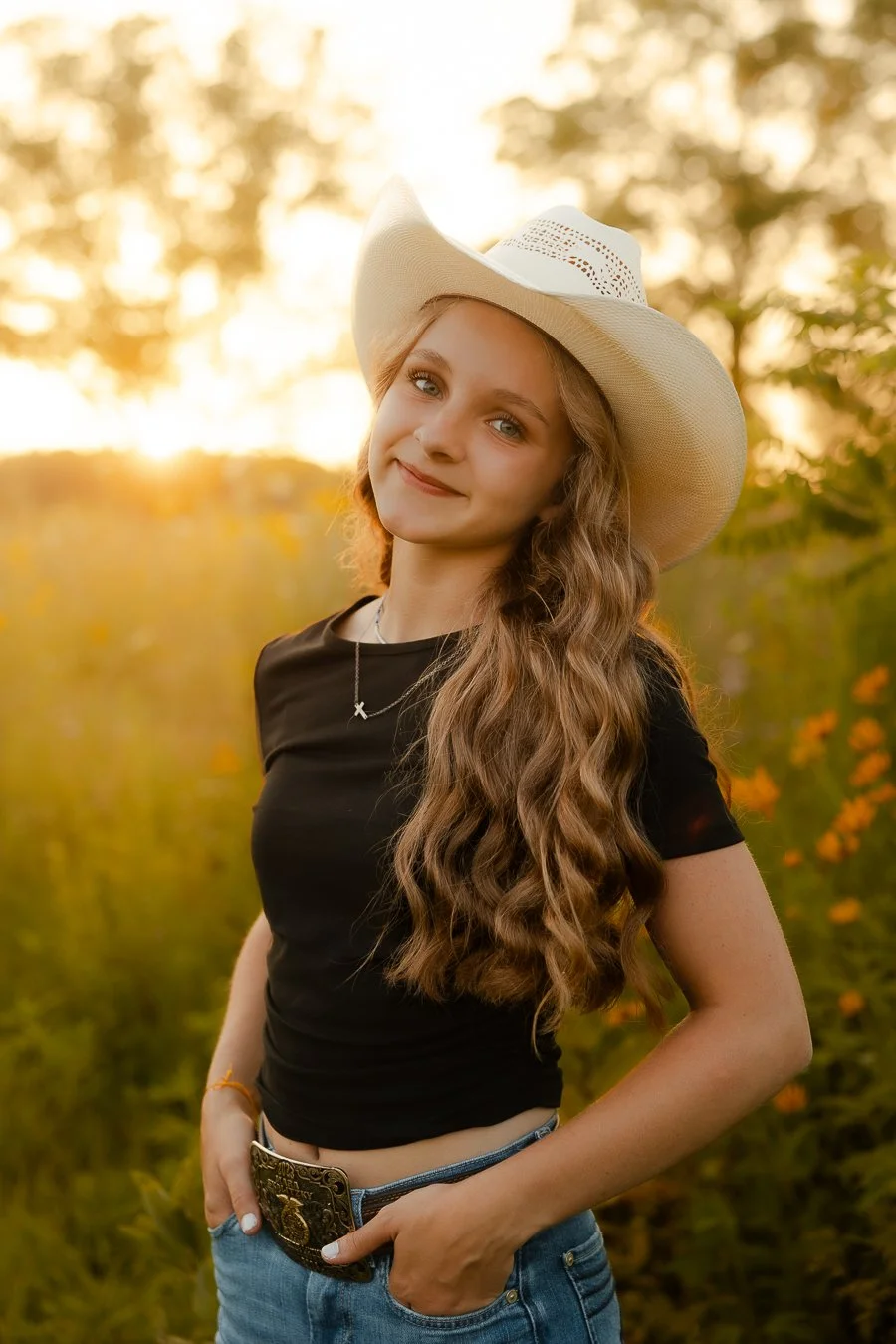 Young woman with long wavy hair wearing a white cowboy hat, black shirt, blue jeans, and a decorative belt outdoors during sunset.