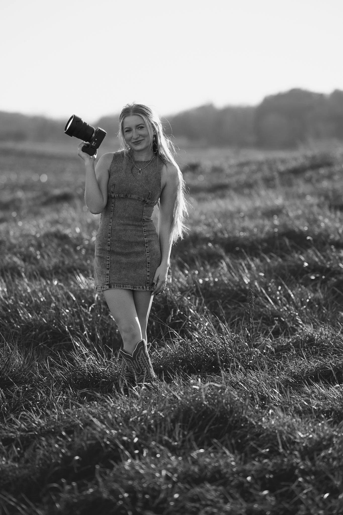 A young woman with long hair in a sleeveless dress and cowboy boots stands in a grassy field, holding a camera in her right hand and smiling.