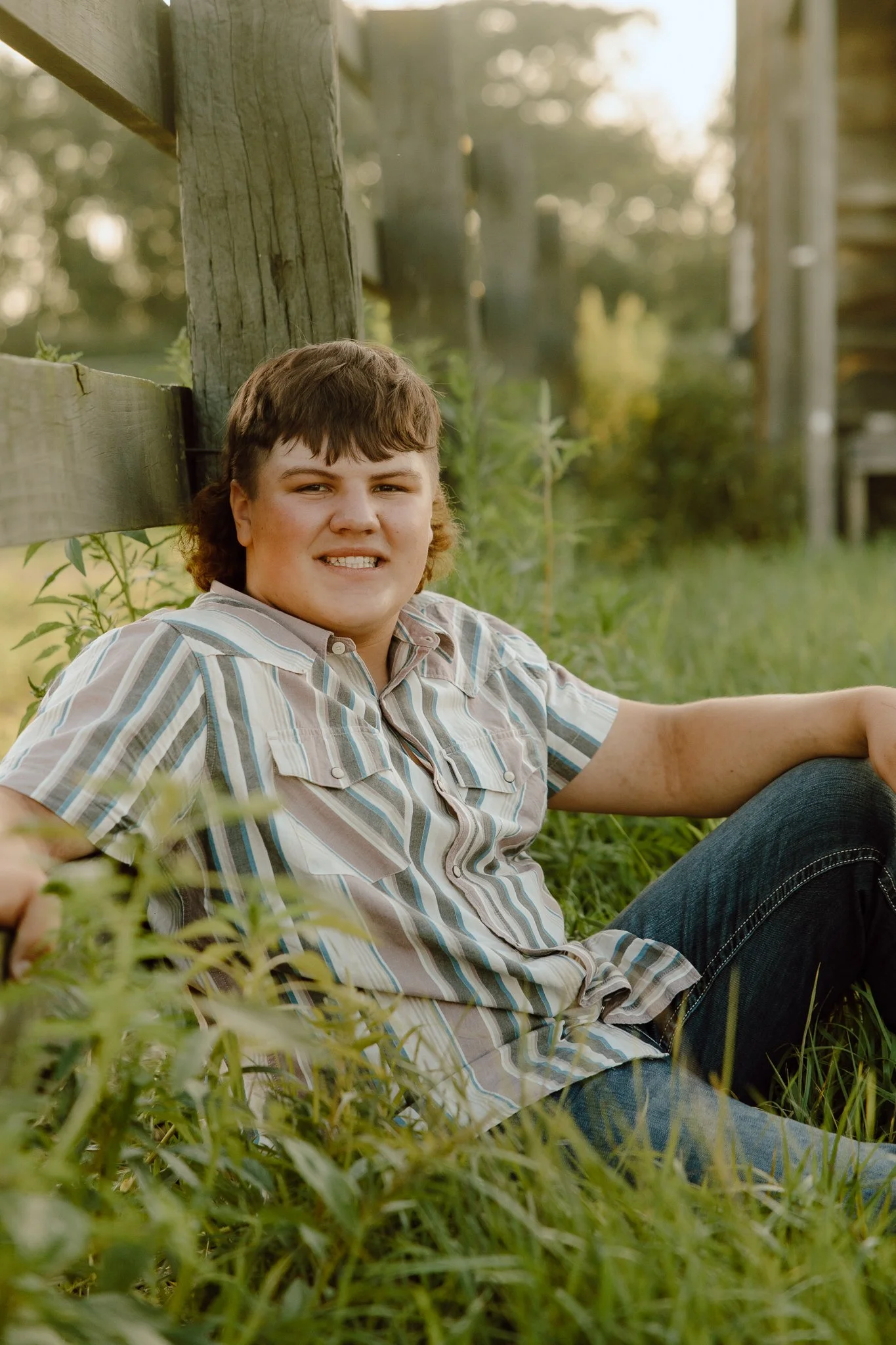 A young person with short brown hair, wearing a striped shirt, sitting on grass near a wooden fence in an outdoor setting, smiling at the camera.
