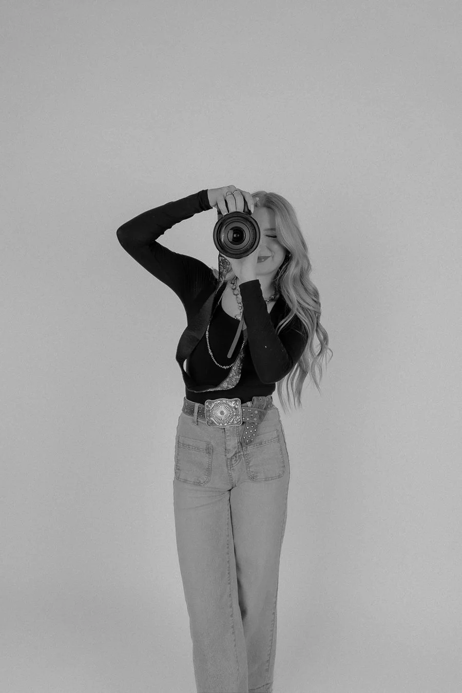 A woman taking a selfie with a camera, smiling, with long wavy hair, wearing a black long sleeve top, high-waisted jeans, and a decorative belt, standing against a plain light-colored background.