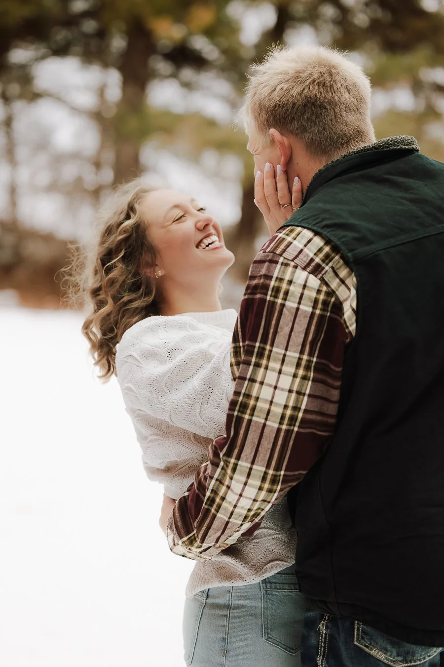 A young woman and a man are outdoors in a snowy area with trees in the background. The woman is smiling joyfully as she touches the man's face, and the man is looking at her with his hands gently on her waist.