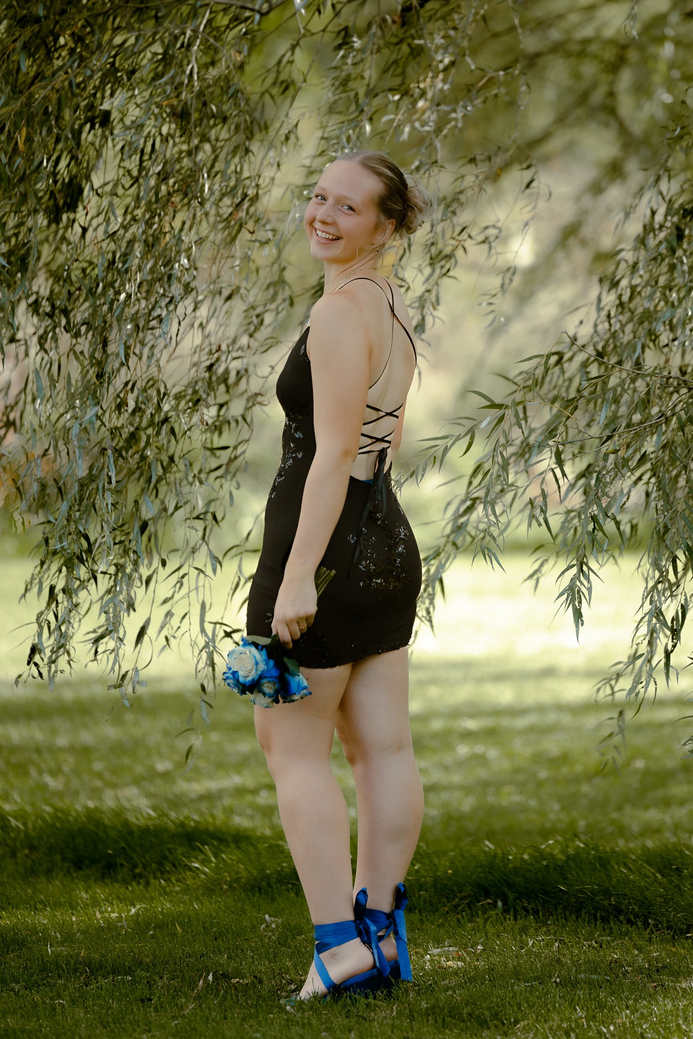A young woman in a black dress with lace-up back, holding a bouquet of blue and white roses, standing outdoors under a tree with green foliage, smiling at the camera.