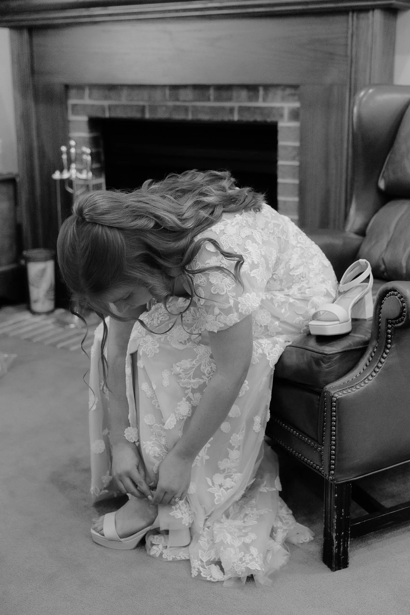 A woman in a lace dress is putting on a high-heeled shoe on her foot while sitting on a chair.